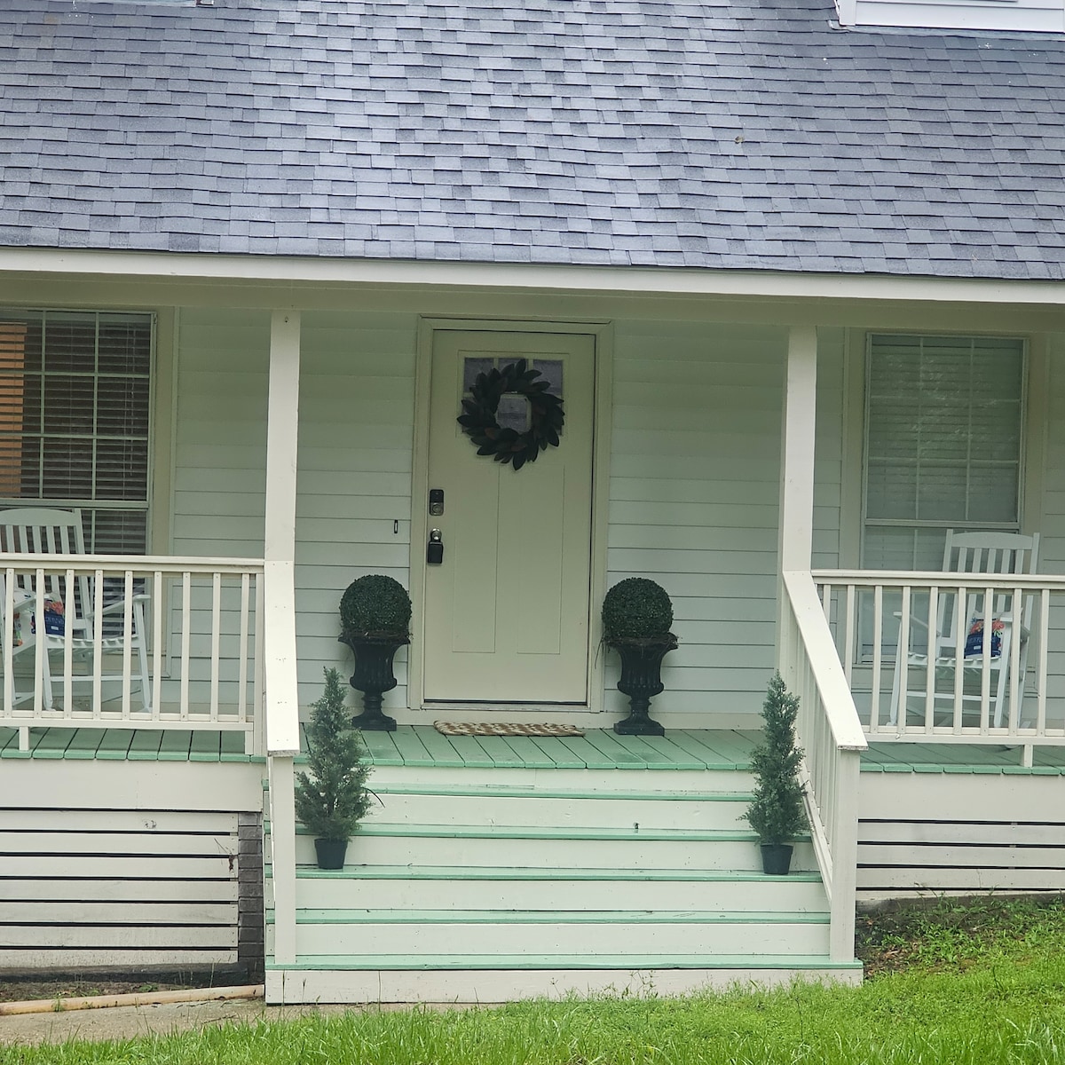 The exterior features a welcoming porch with white railings, two rocking chairs, and a green front door adorned with a wreath. Decorative planters with small evergreen shrubs flank the entrance, enhancing the home's charm. Light green flooring adds a subtle touch to the entryway.