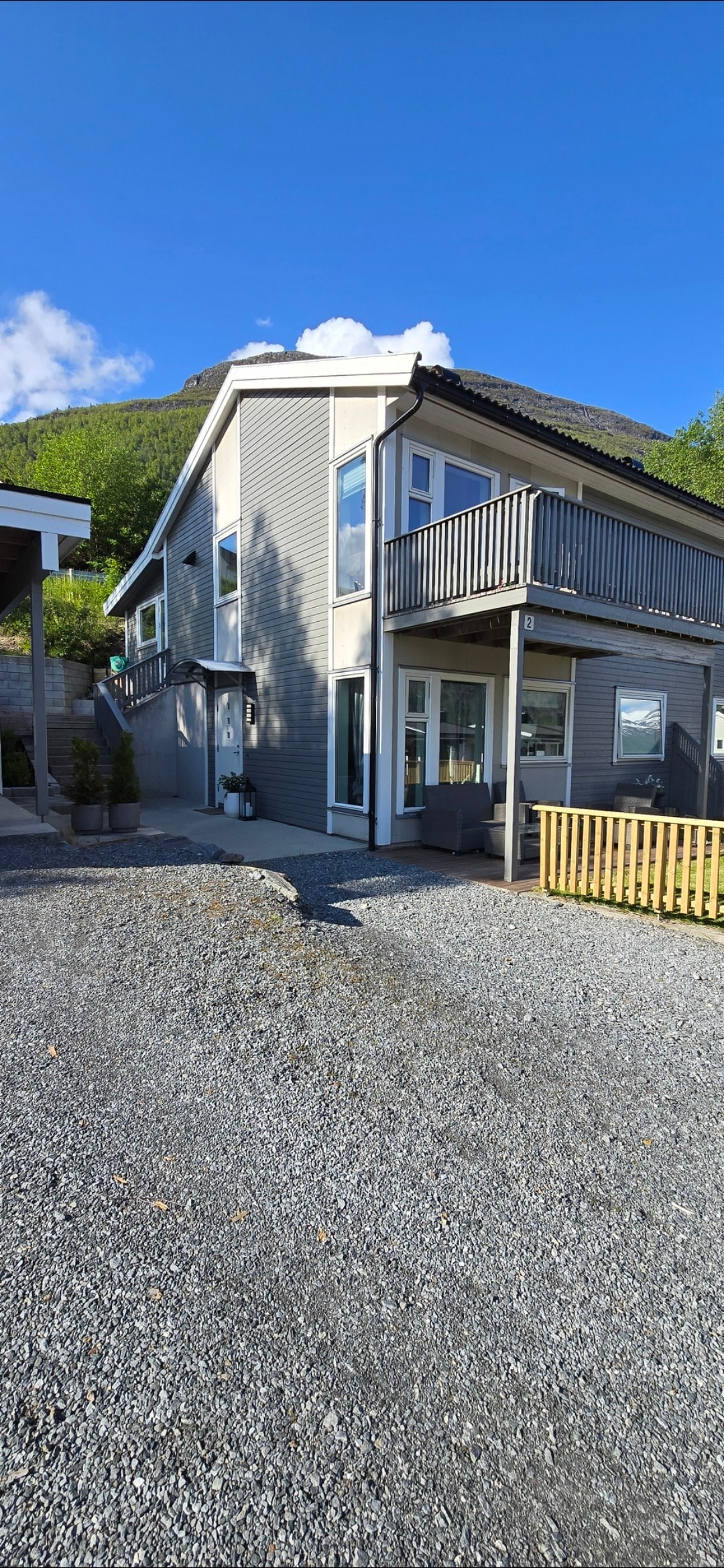 A two-story gray house is framed against a clear blue sky, featuring a balcony adorned with a railing. Windows allow natural light to fill the interior, while a gravel pathway leads to the entrance. Surrounding greenery complements the structure.
