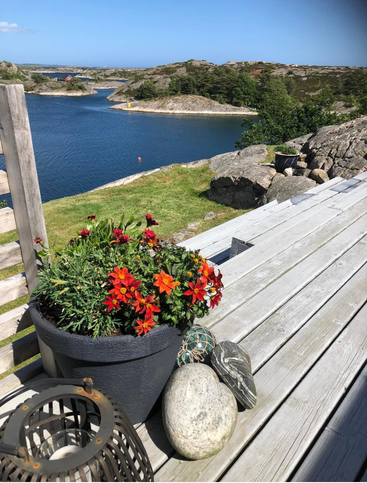 A wooden terrace is seen, adorned with a planter full of vibrant flowers in shades of red and orange. Nearby, smooth stones and a lantern are placed on the surface. In the background, a calm waterfront is visible, reflecting the blue sky and surrounded by rocky landscapes.