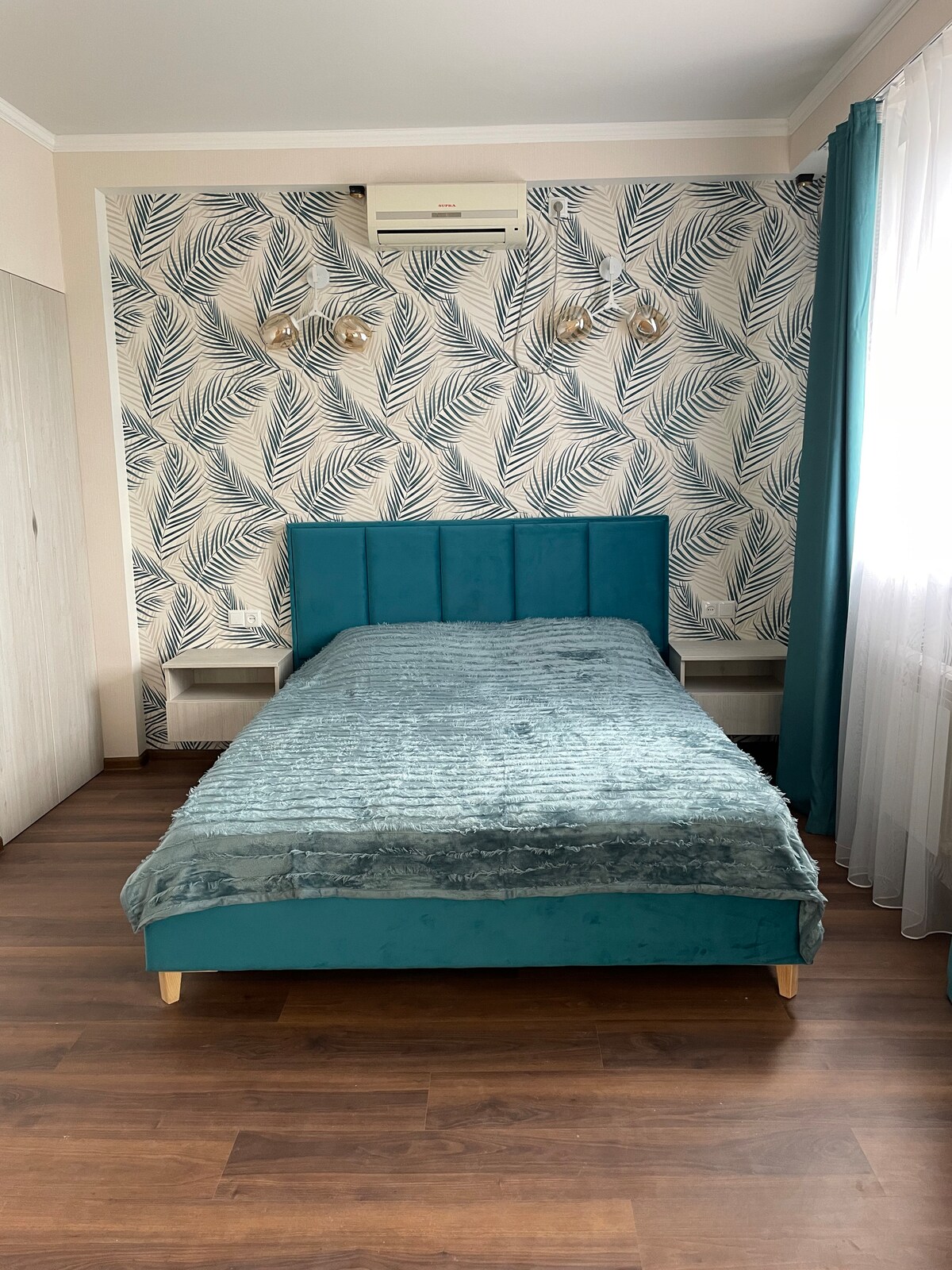 A serene bedroom features a teal upholstered bed, layered with a soft blanket. The walls are adorned with a botanical print, complemented by light and airy curtains. Shelves are positioned beside the bed, providing space for personal items, and an air conditioning unit is mounted above.
