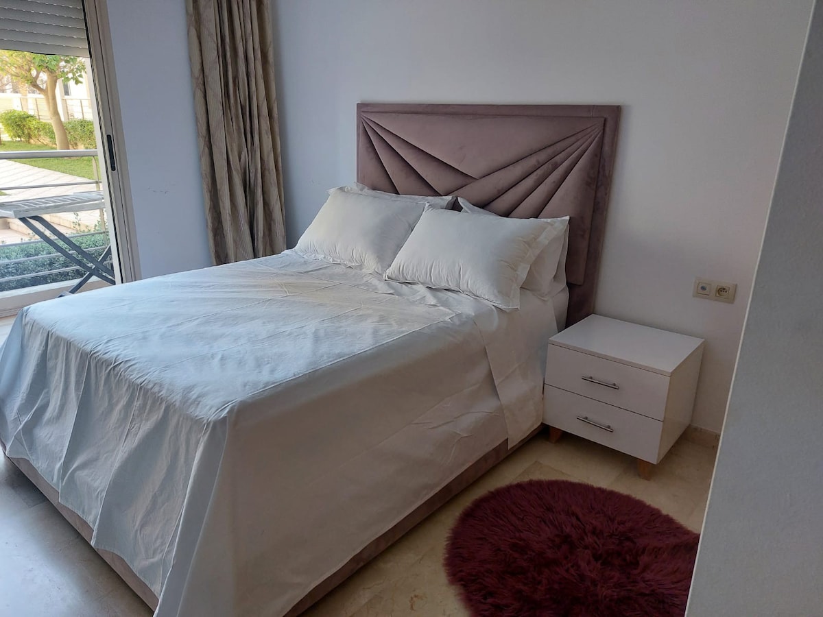 A cozy sleeping area features a spacious bed with a tufted headboard and light bedding. A bedside table sits alongside with a soft rug placed on the floor. Natural light is allowed in through a nearby window, adding a touch of brightness to the room.