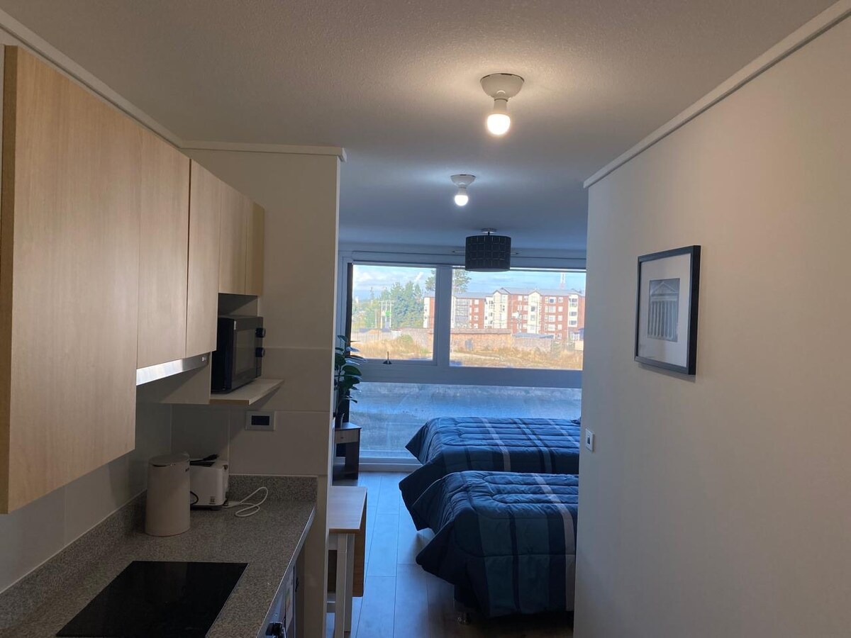 A neat interior space is shown, featuring a kitchenette with light wood cabinetry and a dark countertop. Two spacious beds are positioned in the main area, dressed in blue linens. Large windows provide natural light and a view of surrounding buildings and landscape.