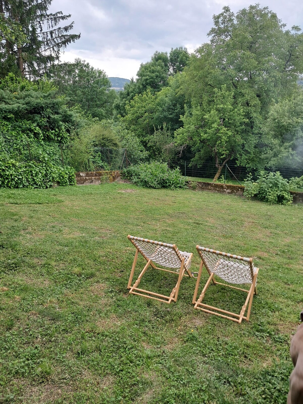 A serene outdoor space is depicted, featuring two comfortable folding chairs positioned on a grassy area. Lush greenery surrounds the space, providing a natural backdrop. A gentle slope of foliage extends into the distance, creating a peaceful setting suitable for relaxation.