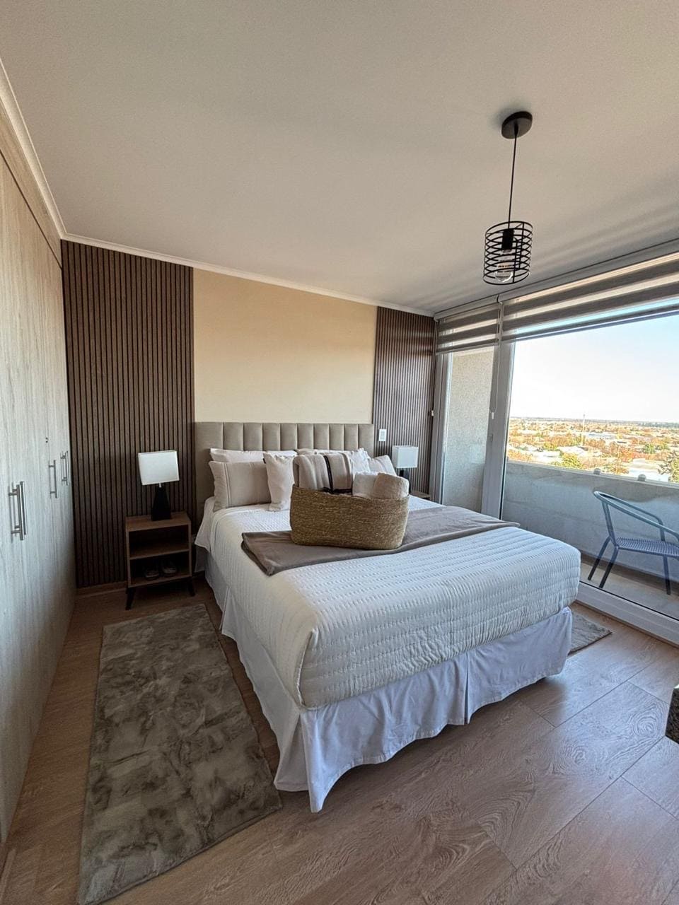 A well-appointed bedroom features a spacious bed with neutral-toned linens and decorative pillows. A bedside table with a lamp complements the modern design. A window allows natural light to flood the space, offering a view of the surrounding area. A small seating area with a chair is also visible.