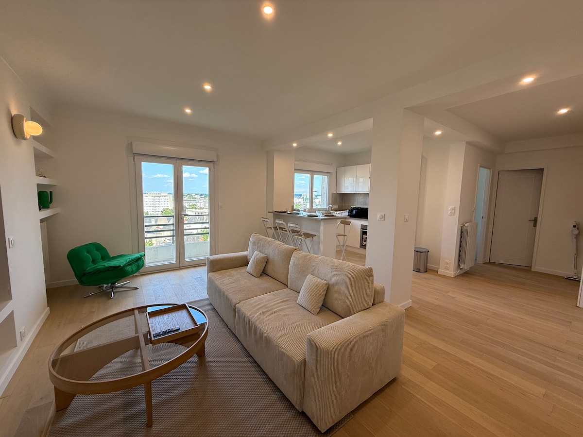 A spacious living area features a light-colored sofa and a wooden coffee table. A comfortable green armchair is positioned nearby. Large glass doors open to a balcony, allowing natural light to fill the space. An open kitchen and dining area can be seen in the background.