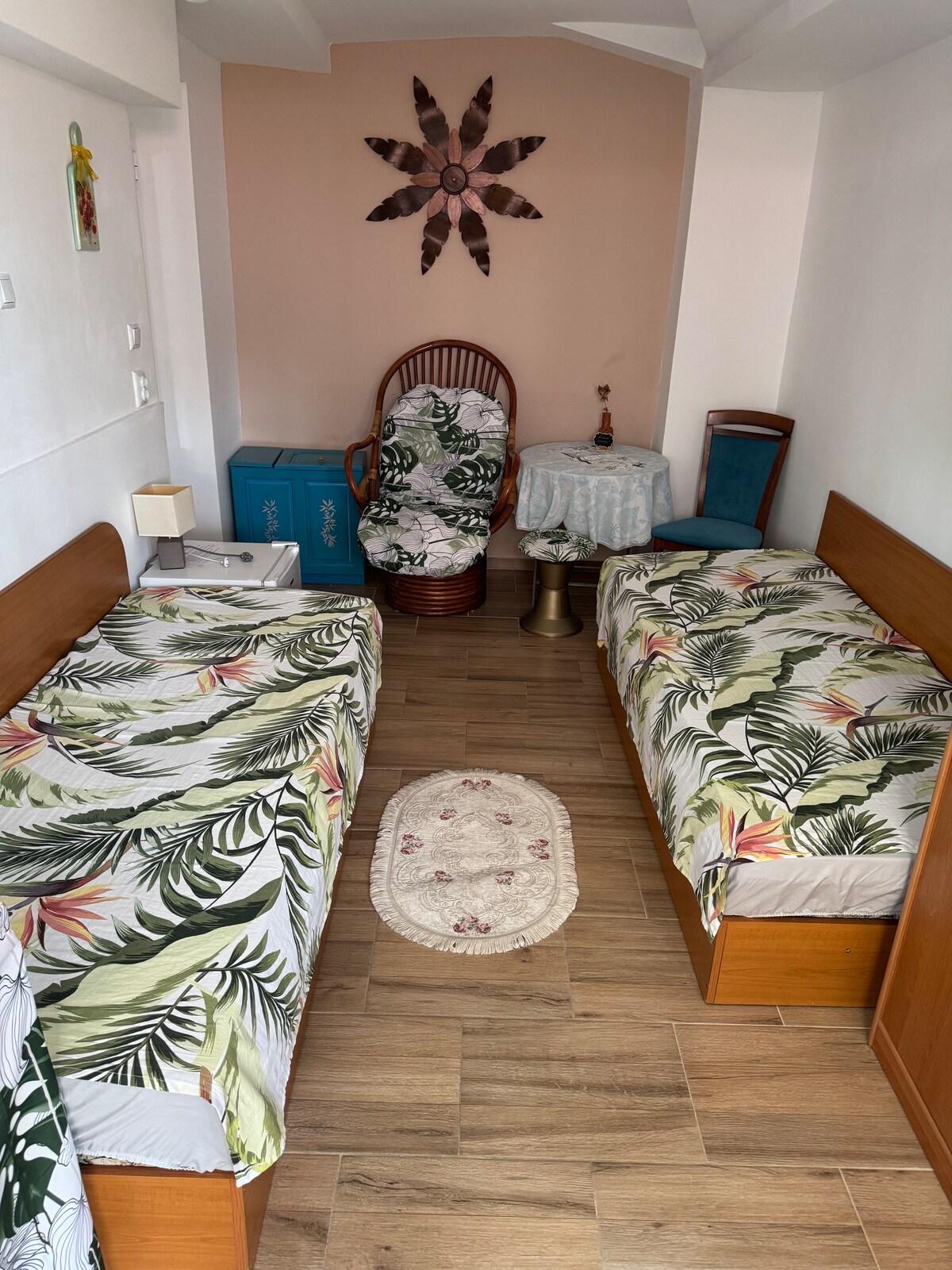 A room featuring two twin beds, each adorned with leafy patterned bed linens. A wicker chair and a small side table are positioned against the wall, with a decorative wall piece above. The floor is made of light wooden planks, contributing to a relaxed atmosphere.