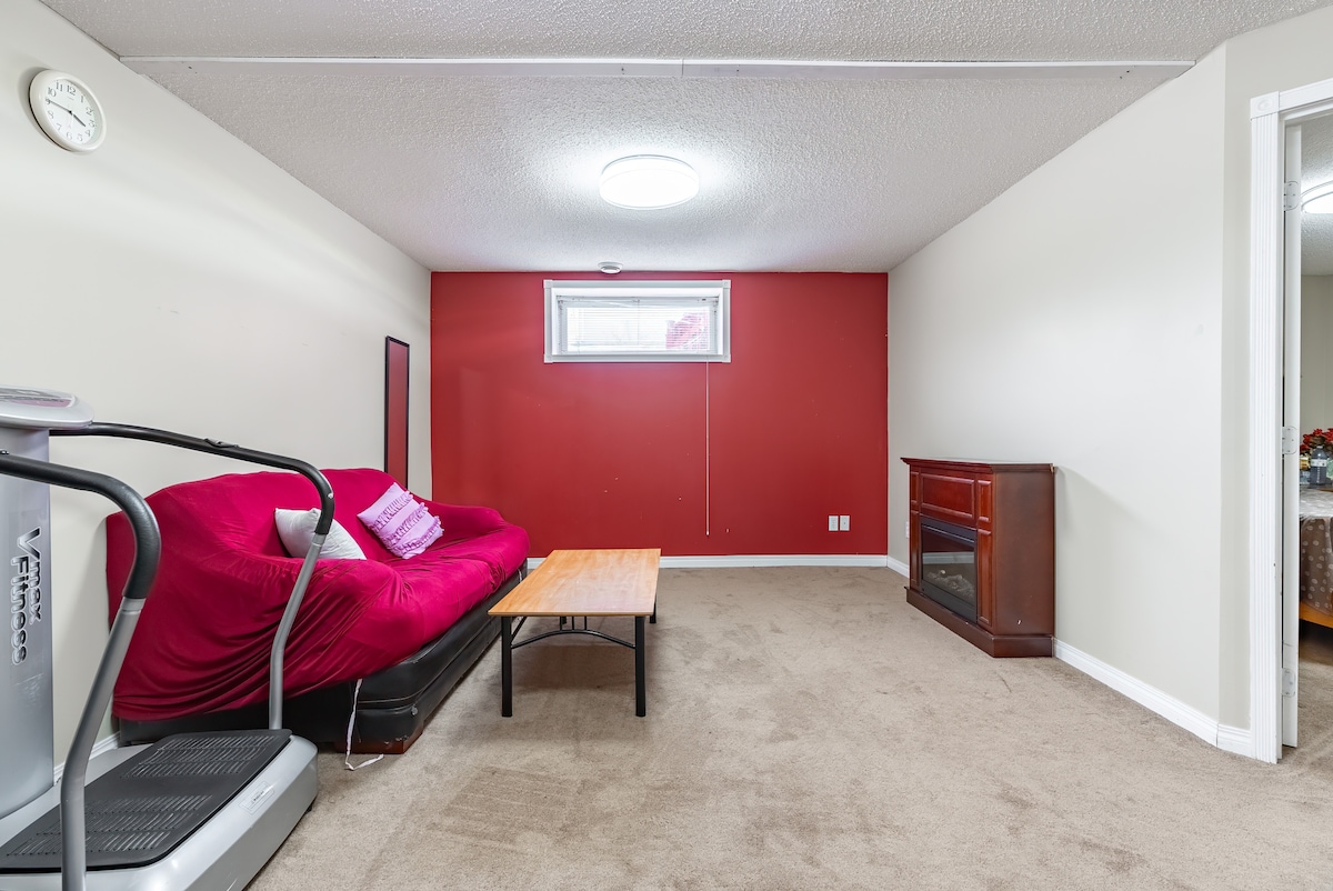 A cozy living area features a red accent wall and neutral-toned carpet. A pink sofa bed is positioned against the wall, accompanied by a wooden coffee table. An electric fireplace stands on one side, with a full-length mirror reflecting the bright, overhead lighting.