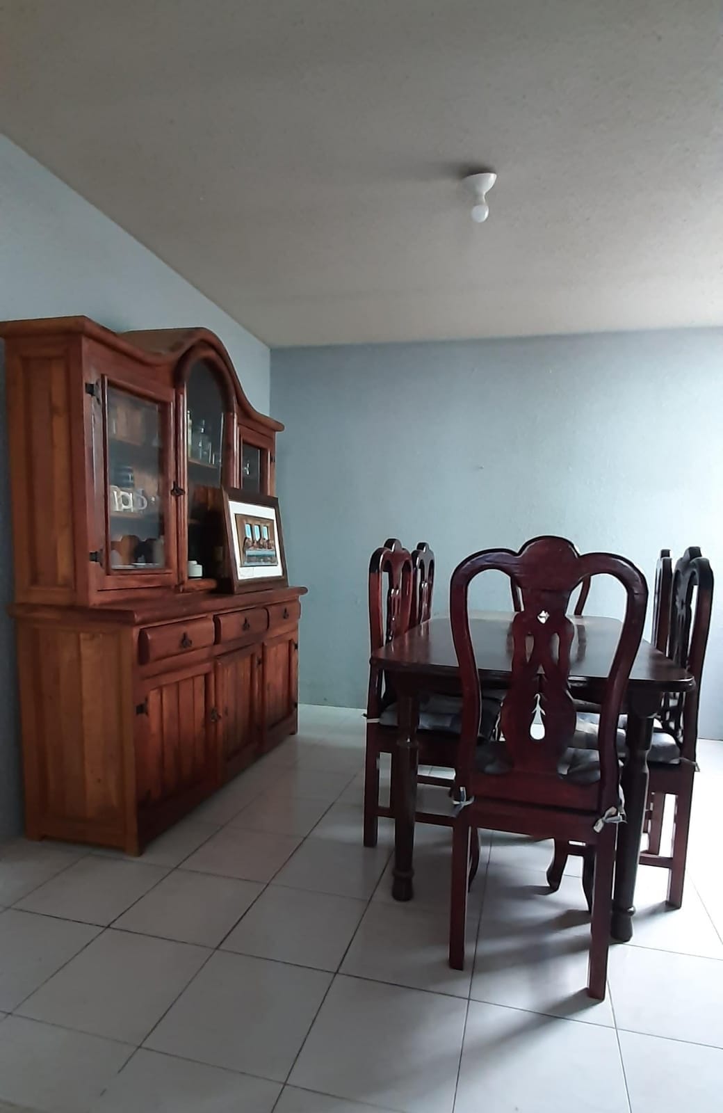 A dining area is visible, featuring a dark wood table surrounded by six chairs. An elegant sideboard complements the space, showcasing glass panels and decorative items. Light enters the room through a nearby window, highlighting the neutral wall color and tiled flooring.