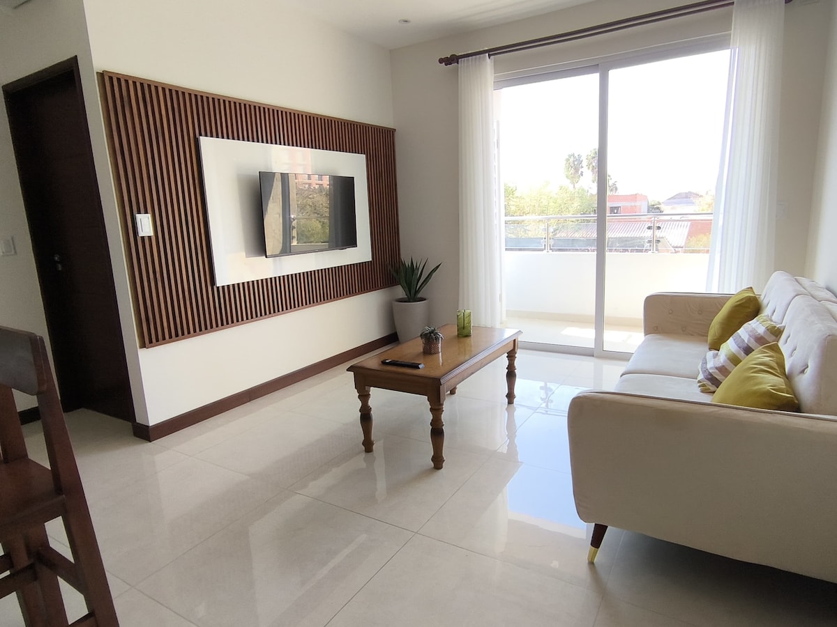A bright living area features a neutral-colored sofa with patterned cushions and a wooden coffee table. A mounted flat-screen TV is positioned against a textured wall. Large windows allow natural light to fill the space, with a balcony view visible in the background.