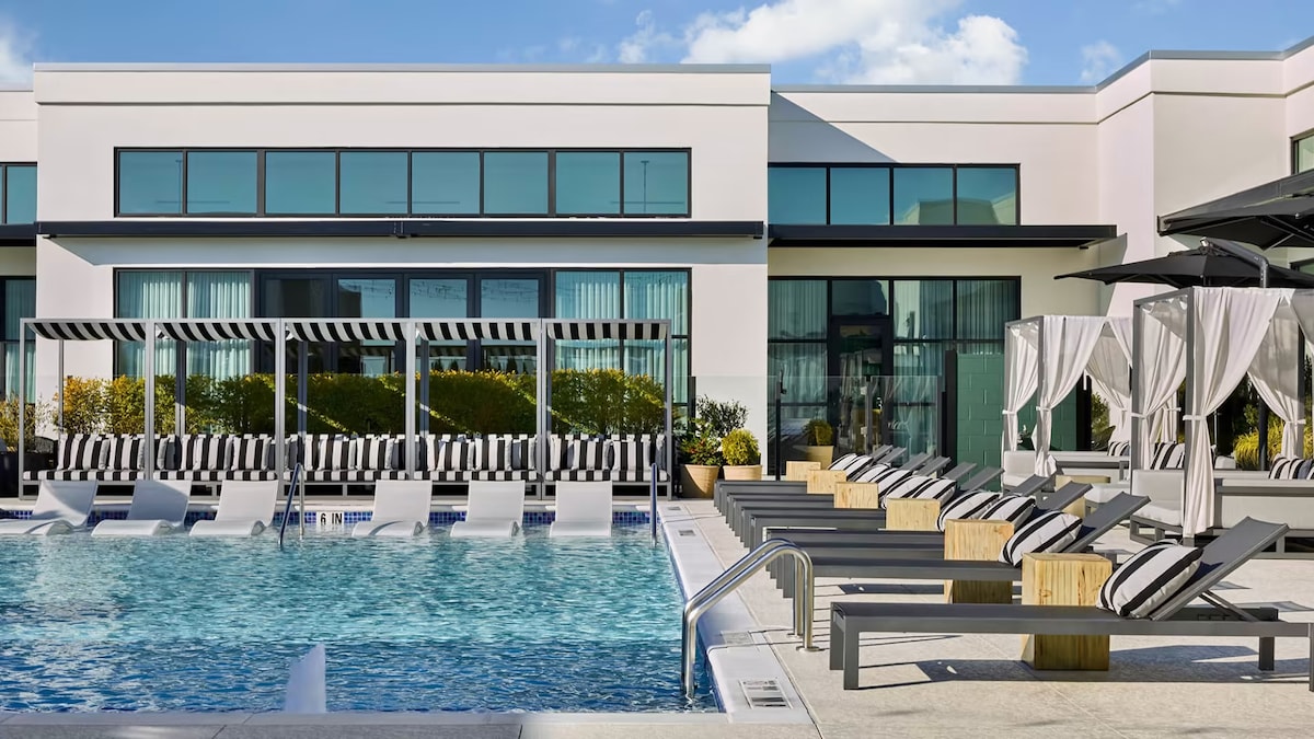 An inviting pool area is shown with lounge chairs arranged around the water's edge. Stylish cabanas provide shaded seating options. Large glass windows reflect natural light from the buildings nearby, enhancing the vibrant atmosphere of the space.