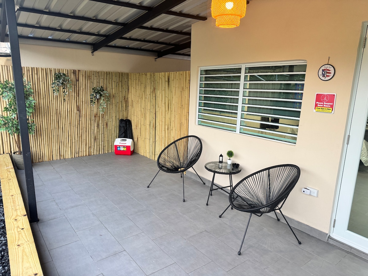 A serene balcony space is featured, furnished with two black accent chairs and a small table. A soft glow is provided by an overhead light. Bamboo-style wall panels and potted plants enhance the environment, creating a comfortable area for relaxation.