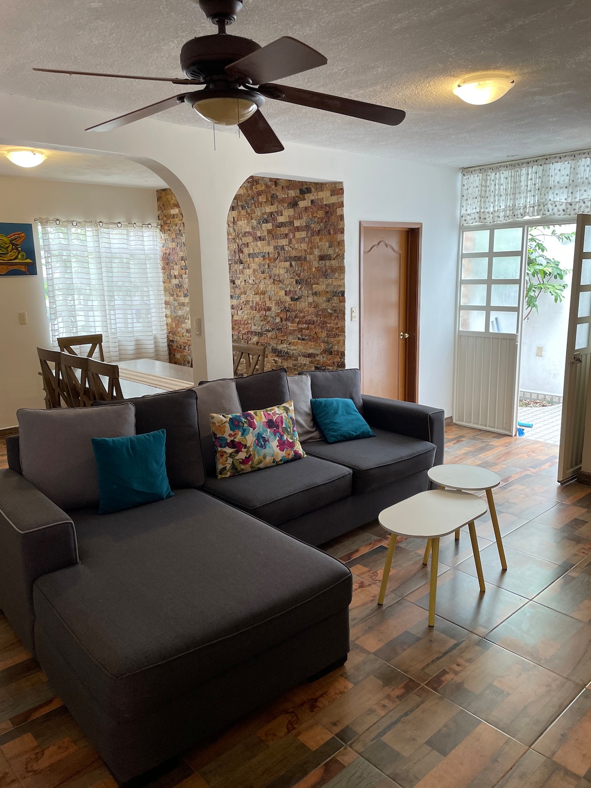 A spacious living area features a gray sectional sofa adorned with colorful cushions. Light streams through the large windows, illuminating the room's warm tones. A neutral-toned wall contrasts with a textured brick accent, while two small side tables are positioned next to the sofa.