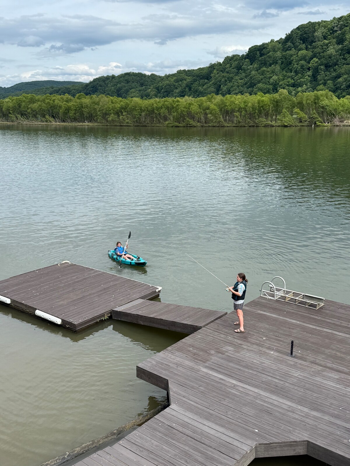 A wooden dock extends into a calm body of water, where a person is fishing while another kayaks nearby. Lush greenery can be seen along the water's edge, with hills rising in the background under a partly cloudy sky.