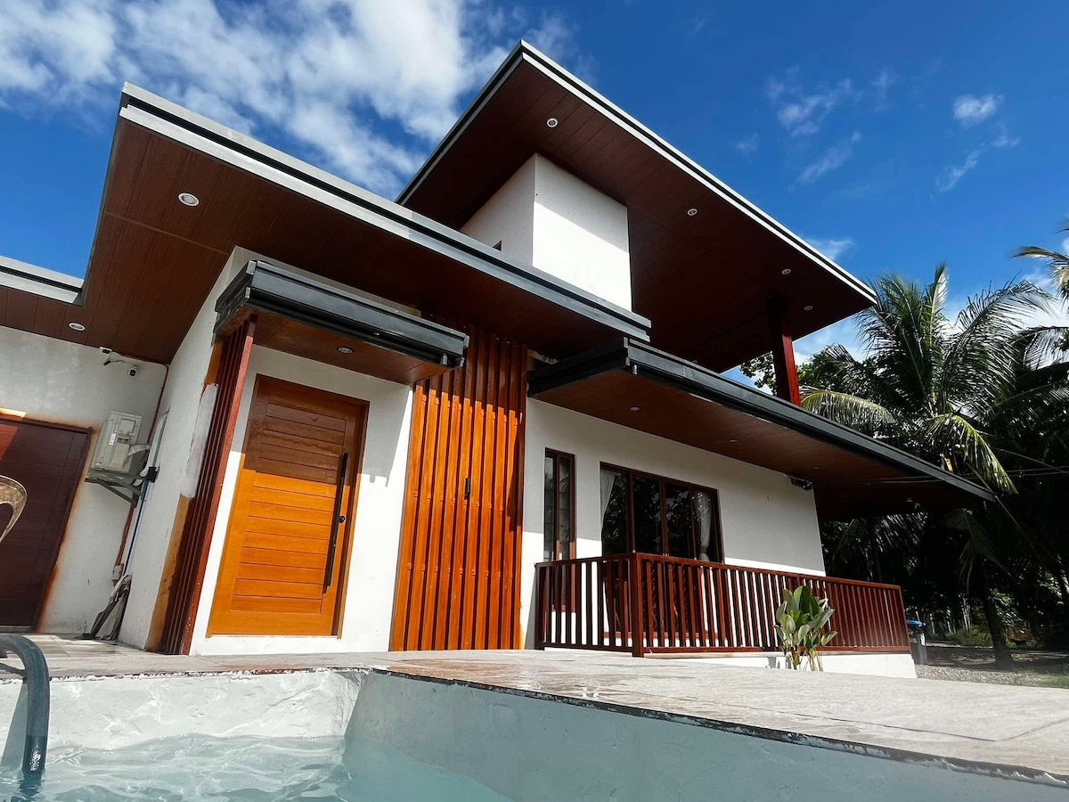 A modern house features a combination of wood and white exterior, with large windows providing natural light. The entrance showcases a wooden door. Lush greenery and palm trees surround the property, enhancing its tranquil setting. A swimming pool is positioned in the foreground.