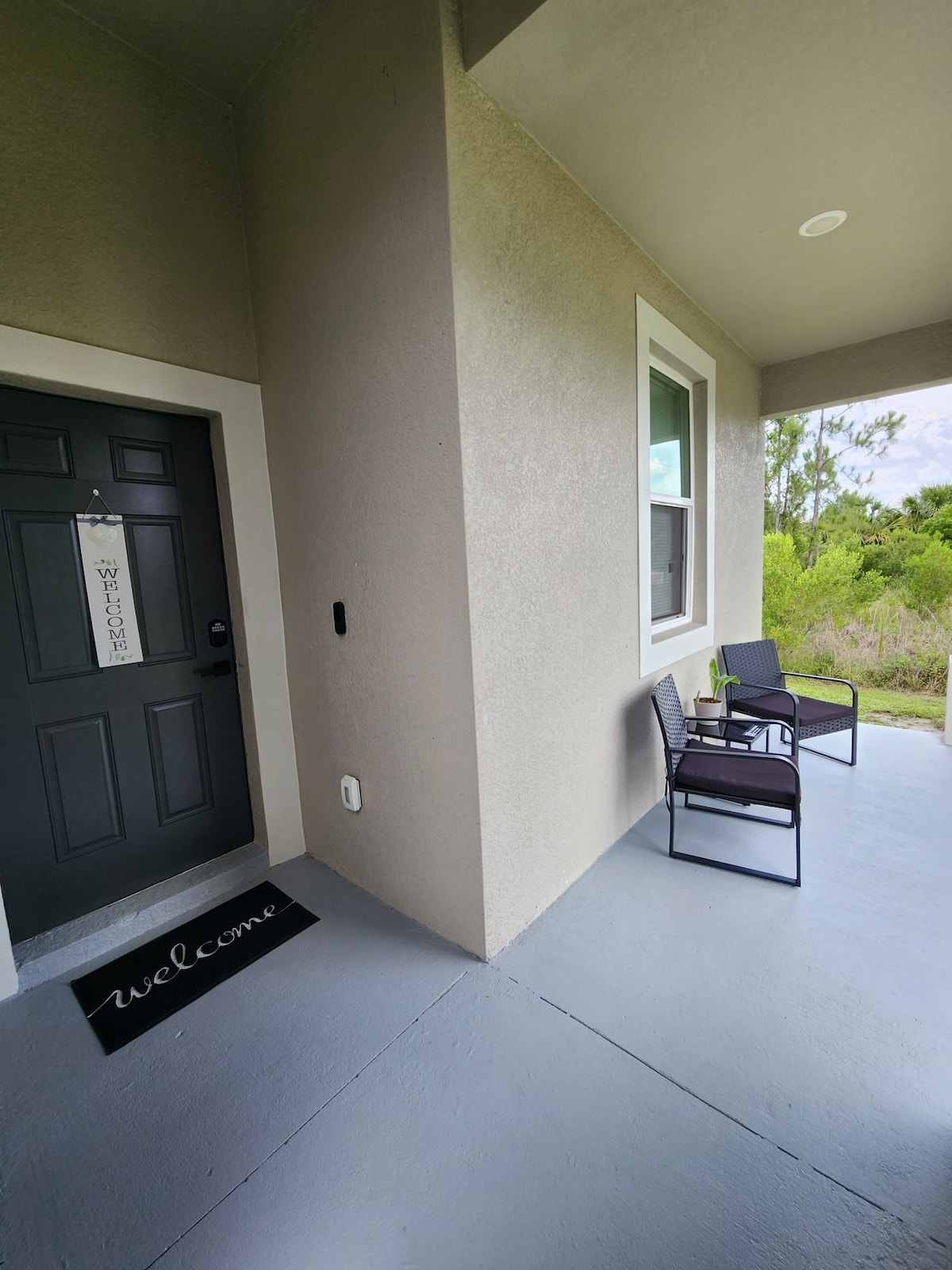 An inviting front porch features a dark door with a 'welcome' sign. Two black chairs are positioned beside the entrance, complemented by a simple welcome mat on the gray floor. Lush greenery is visible in the background, enhancing the tranquil setting.