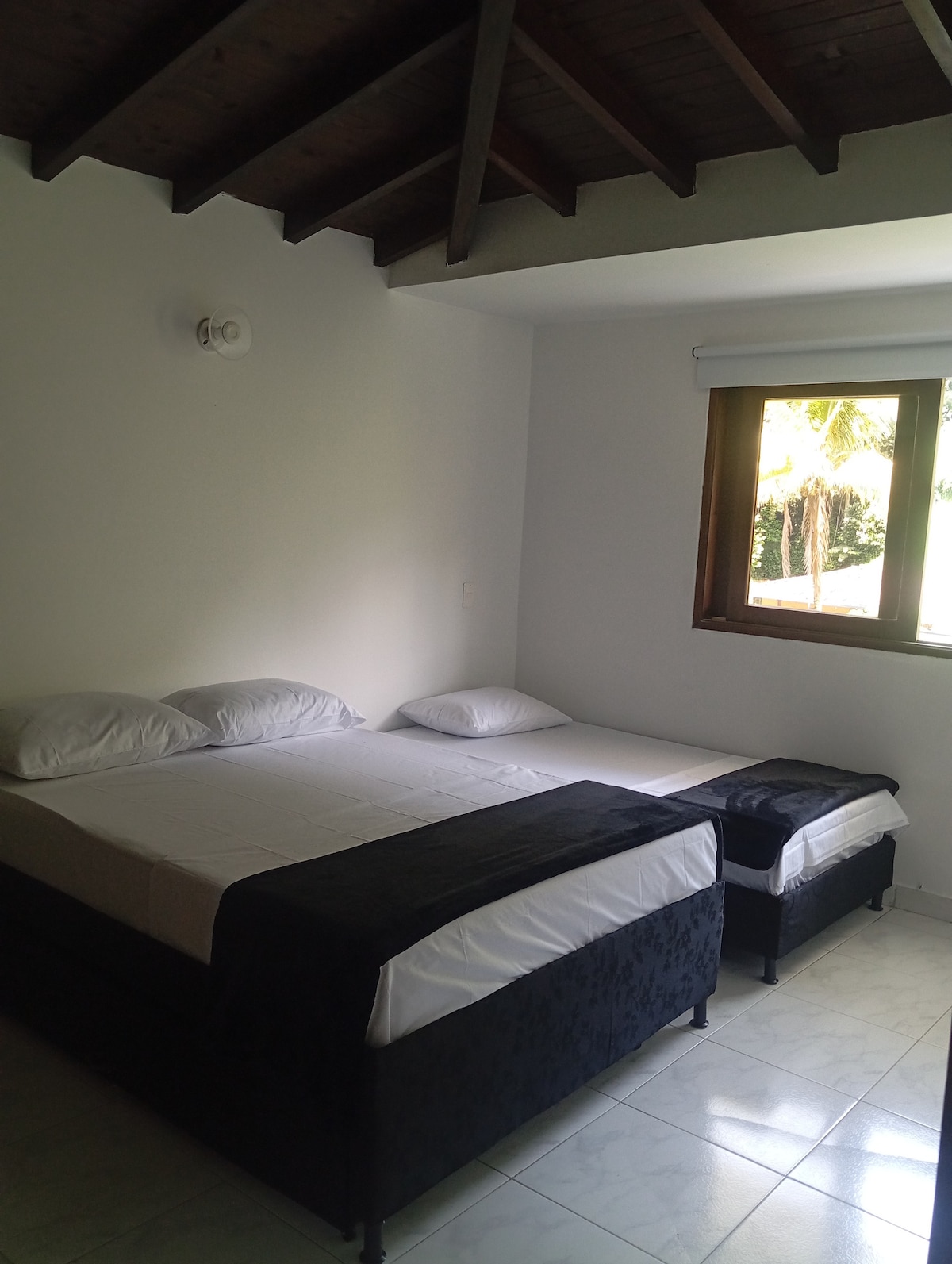 A well-lit bedroom is shown, featuring two spacious beds. One bed is framed in dark upholstery, while the other is a twin-sized bed positioned beside it. Natural light enters through a window, illuminating the clean, tiled floor and the simple decor.
