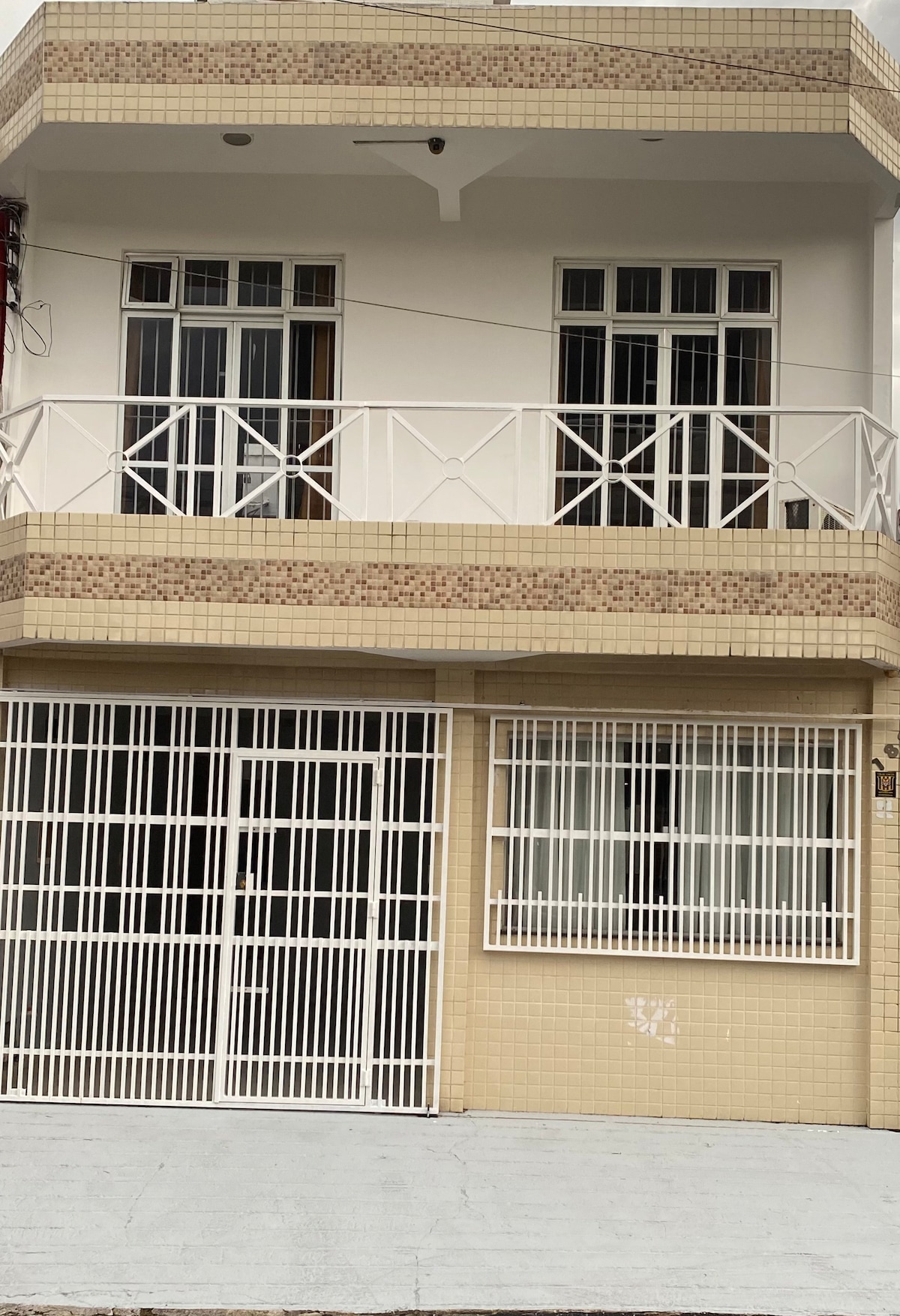 The exterior of a two-story house is visible, featuring a tiled facade with decorative elements. A gated area is situated at the bottom, and two balconies with white railings are positioned on the upper level. Clear glass windows provide a view of the interior.