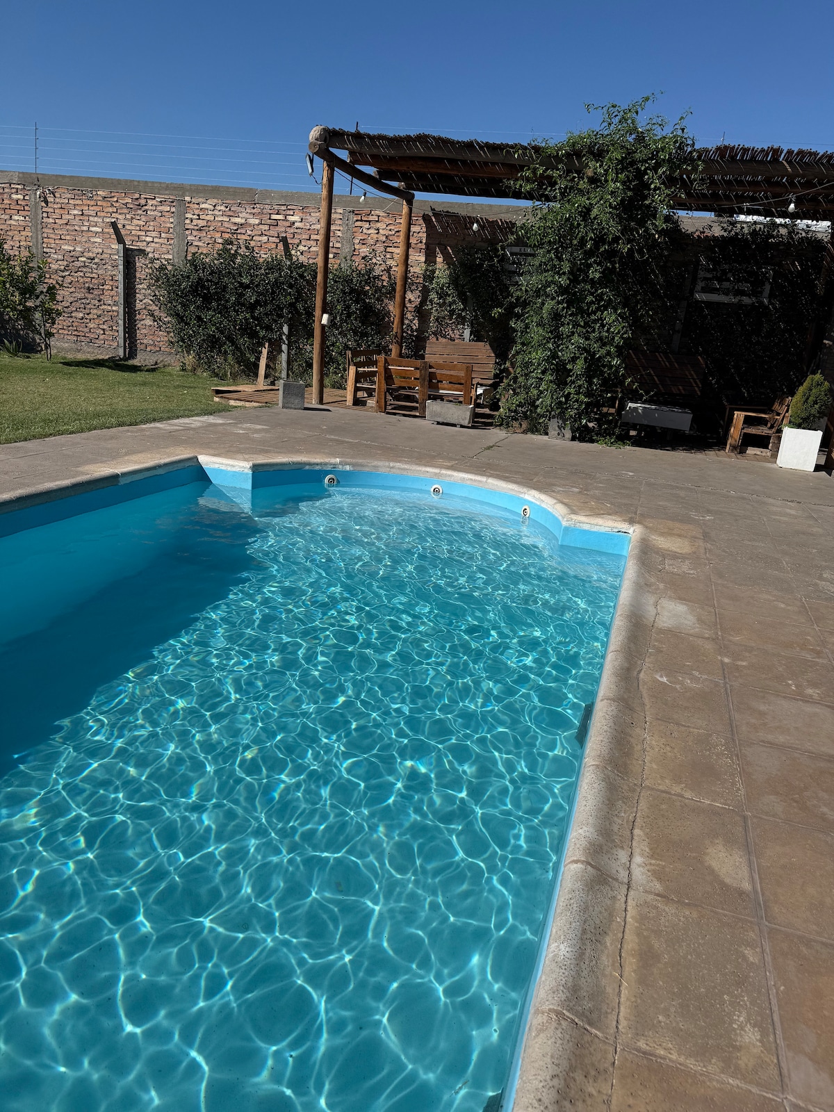 A refreshing swimming pool is featured, surrounded by paved stone decking. Lush greenery and a shaded seating area with wooden chairs are visible in the background, offering a tranquil outdoor space. Clear blue water glistens under sunlight, inviting relaxation.
