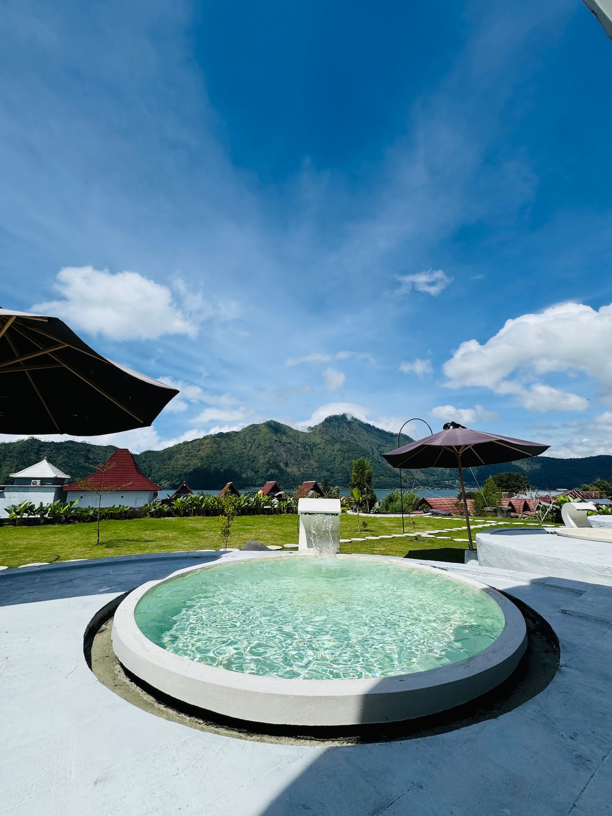 A hot tub is situated outdoors, surrounded by vibrant green grass and nestled against a backdrop of mountains. Two dark umbrellas provide shade, creating a relaxing atmosphere under a clear blue sky filled with fluffy white clouds.