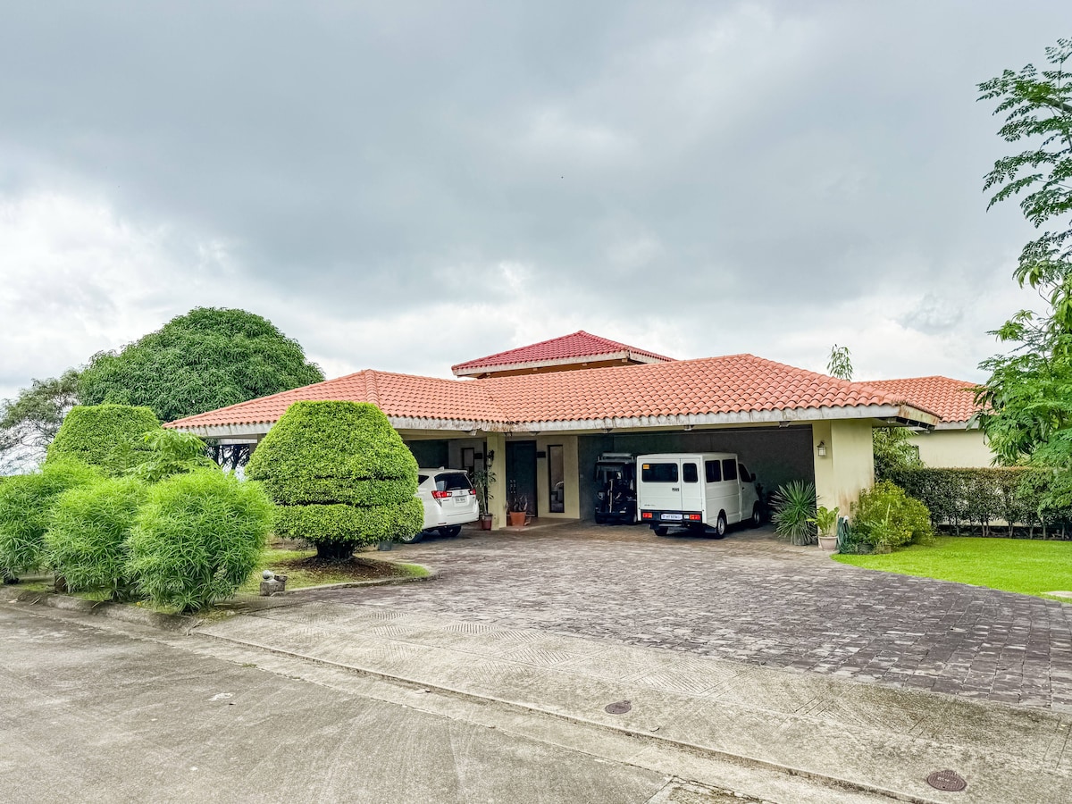 The exterior of the guest house is displayed, featuring a broad driveway with two parked vehicles. A terracotta roof complements the structure, surrounded by well-manicured greenery and shrubs. The landscape presents a serene atmosphere, showing a glimpse of the inviting entrance.
