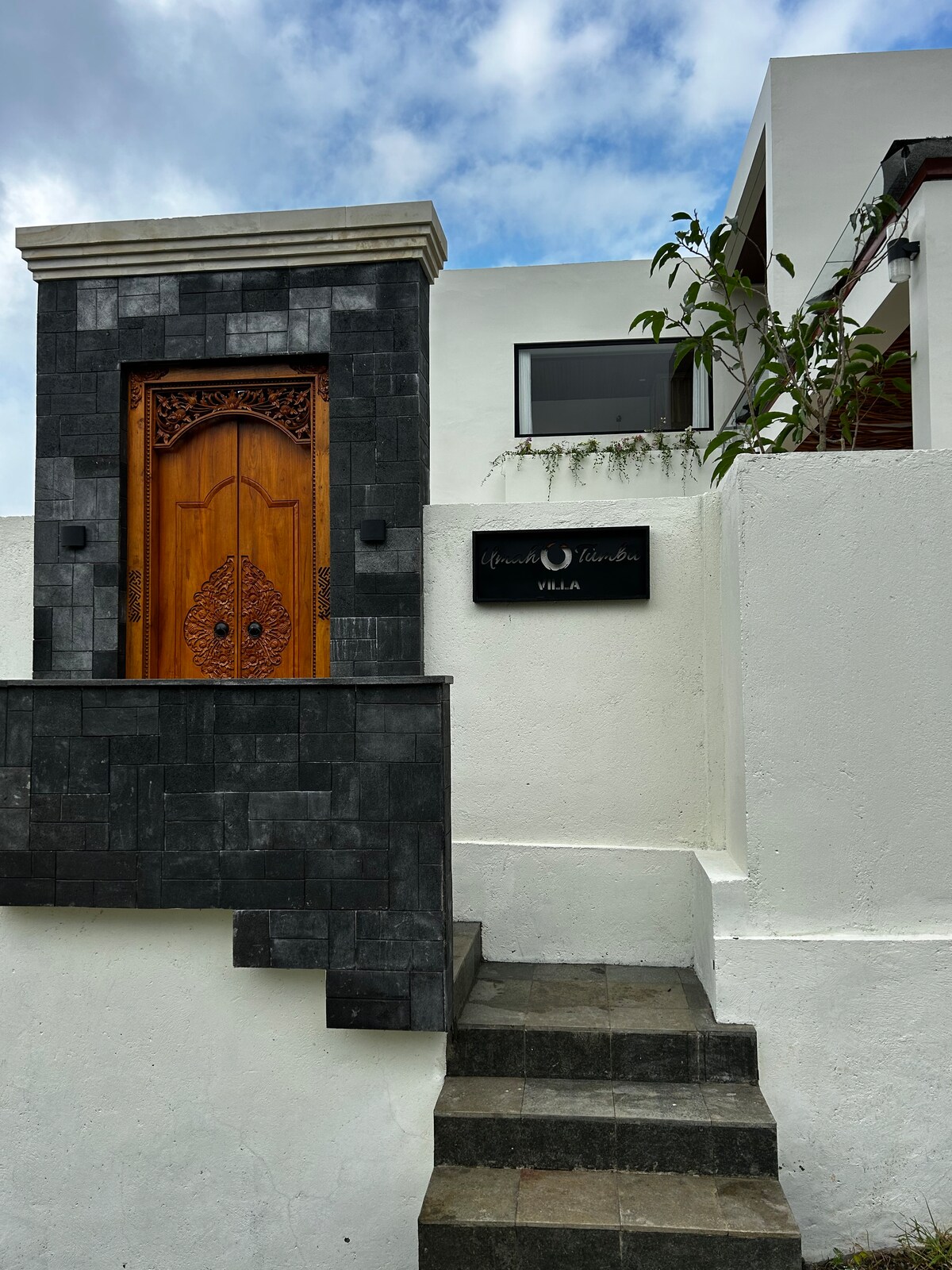 A decorative wooden door with intricate carvings is framed by a stone entrance, leading to the property. The structure features a combination of dark stone and white walls. A sign is displayed beside the door, indicating the name of the villa. Green foliage adds a touch of nature.