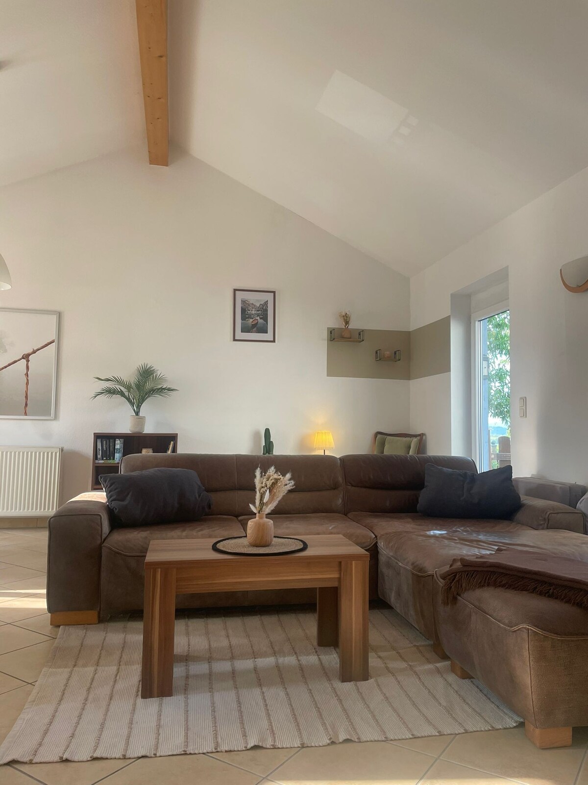 A spacious living area features a sectional sofa with soft brown upholstery and a wooden coffee table at its center. An area rug lies beneath, while natural light streams in through large windows. Simple decor elements are present, including a plant and framed artwork on the walls.
