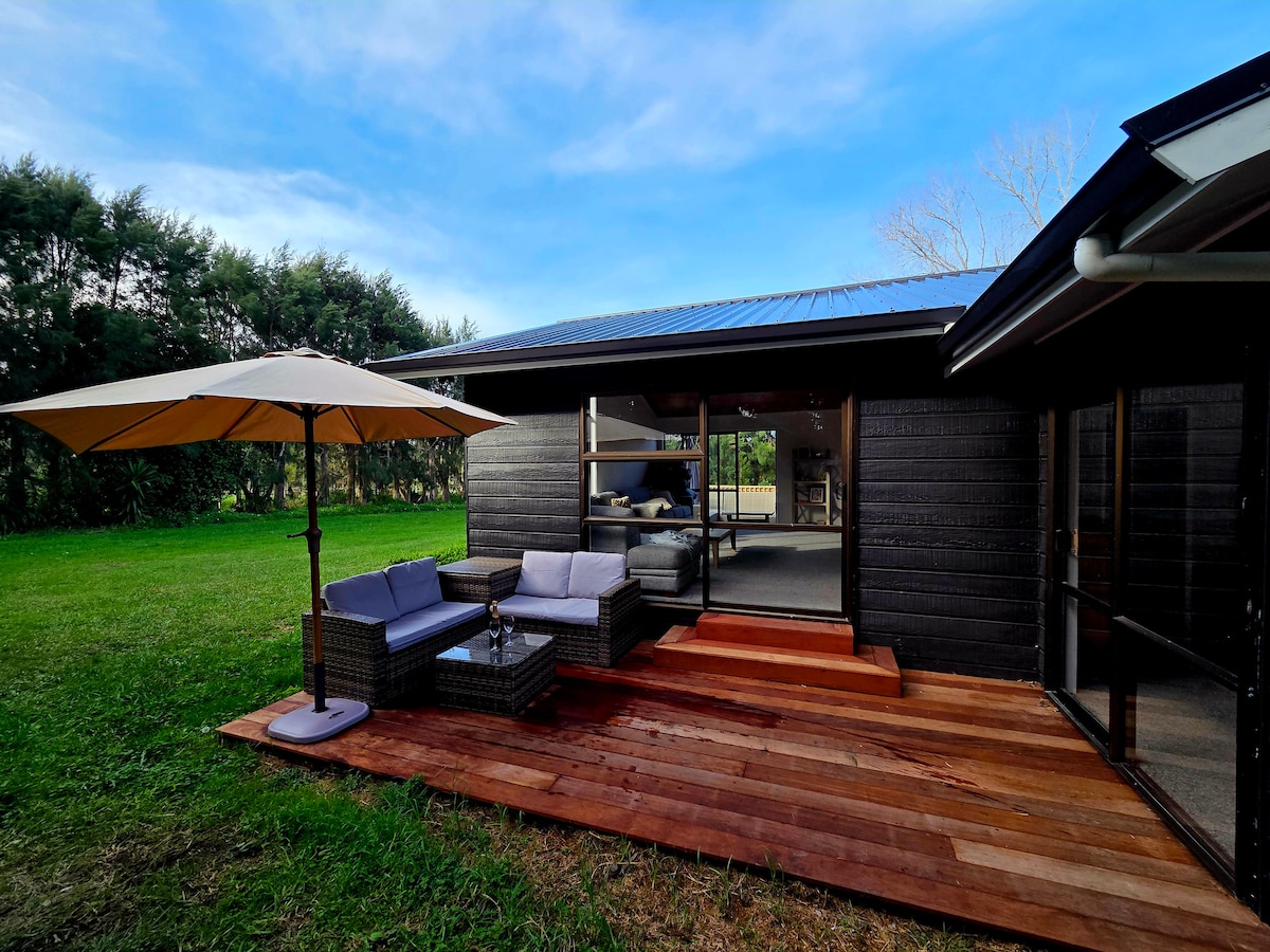 An exterior view of the home reveals a wooden deck with comfortable seating, shaded by a large umbrella. Lush green grass surrounds the deck, and large glass doors provide access to the interior space, inviting natural light and creating a seamless transition between indoor and outdoor areas.