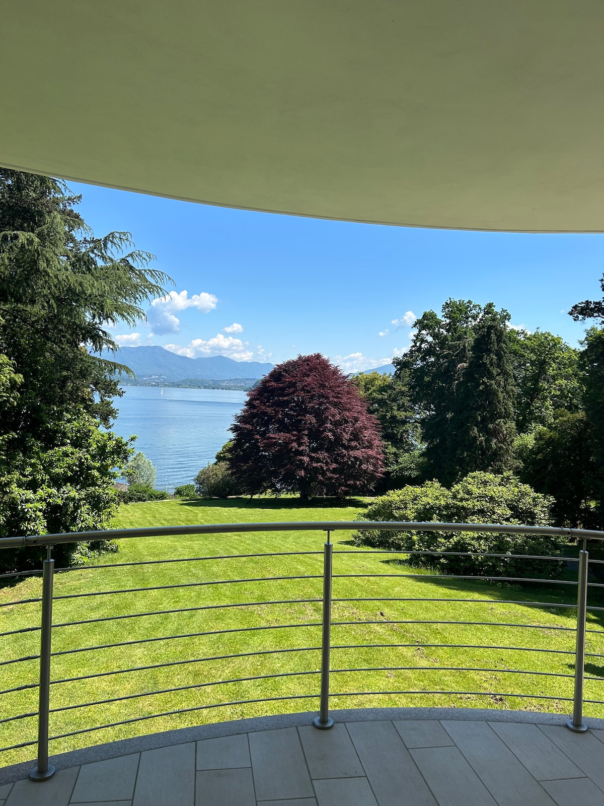 A spacious terrace overlooks the lush garden and serene Lake Maggiore. A vibrant tree stands prominently against the backdrop of rolling hills and clear blue skies, while well-maintained grass extends towards the water's edge, creating a peaceful outdoor environment.