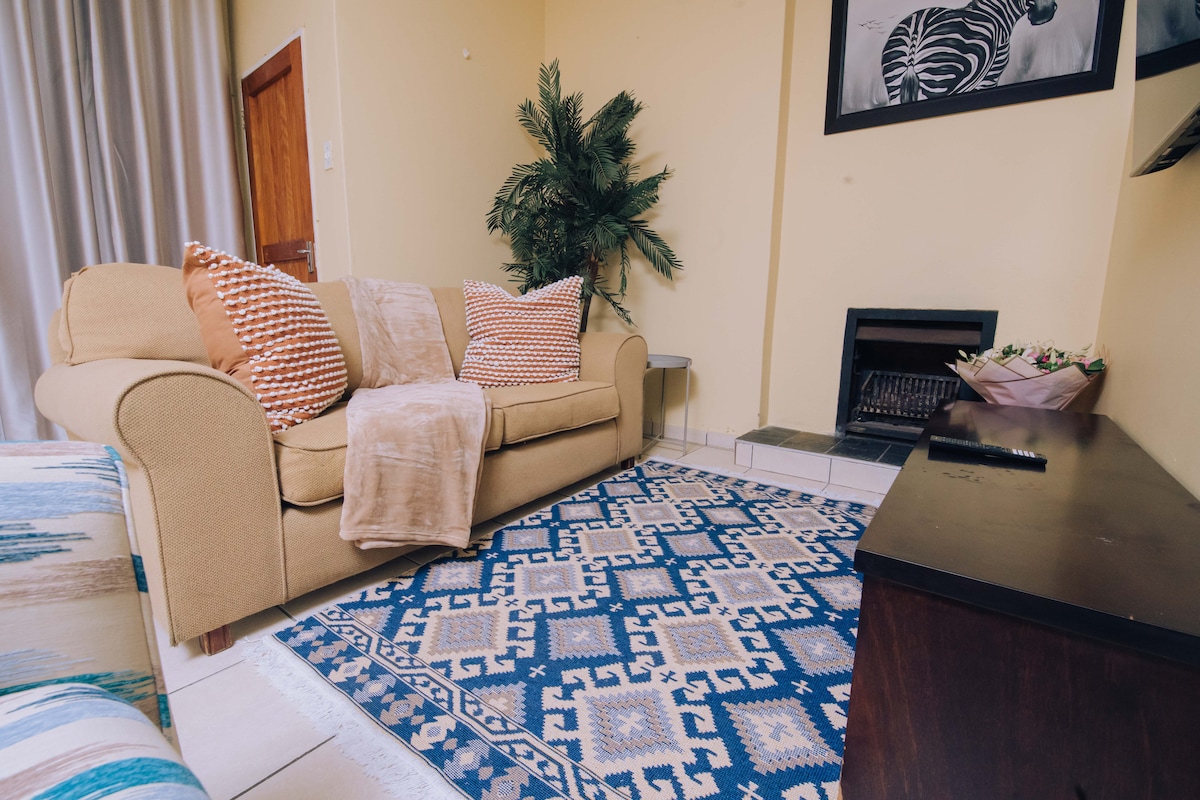 A cozy living area features a light-colored sofa adorned with patterned cushions, complemented by a soft throw. A decorative area rug with geometric patterns anchors the space, while a small indoor plant adds a touch of greenery. A fireplace is positioned near the wall.
