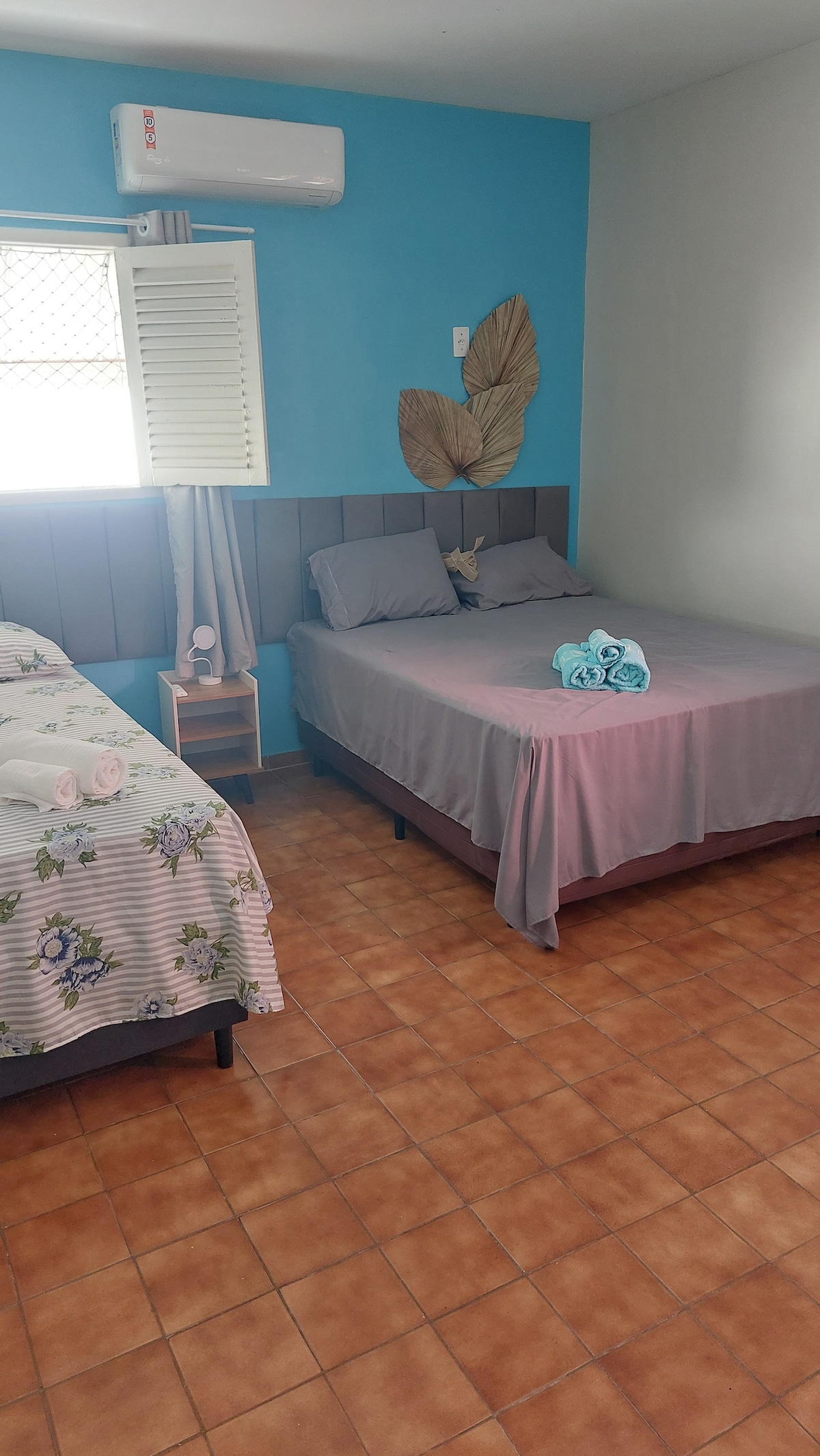 A bright and comfortable room features two beds with simple, modern bedding. A decorative wall is accentuated by large leaf motifs. A small nightstand stands beside one of the beds. Light-colored tiled flooring adds to the room's inviting feel, complemented by natural light from the window.
