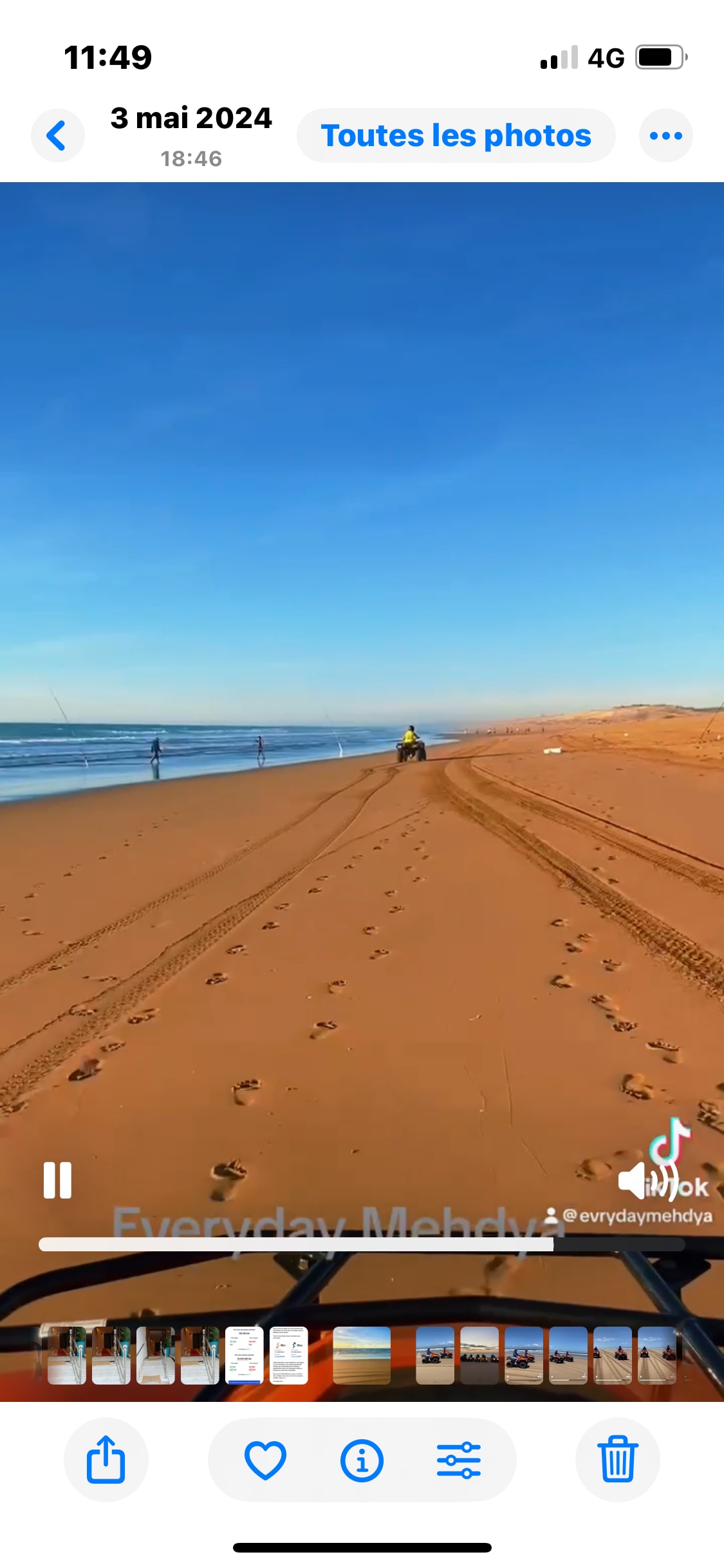 A serene beach scene is captured under a clear blue sky, with gentle waves lapping at the shore. Footprints are visible in the warm, sandy terrain, leading towards a small vehicle and a few distant figures enjoying leisure activities along the water's edge.