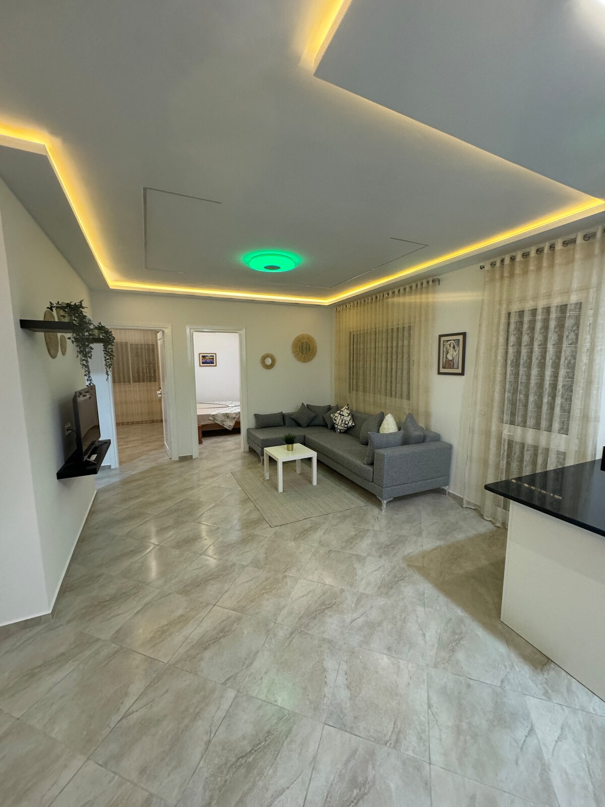 A living area features a comfortable gray sofa with decorative cushions, positioned beside a small white table. Soft lighting emanates from the ceiling, enhancing the space. Large windows are dressed with sheer curtains, allowing natural light to filter in, while a flat-screen TV is mounted on the wall.