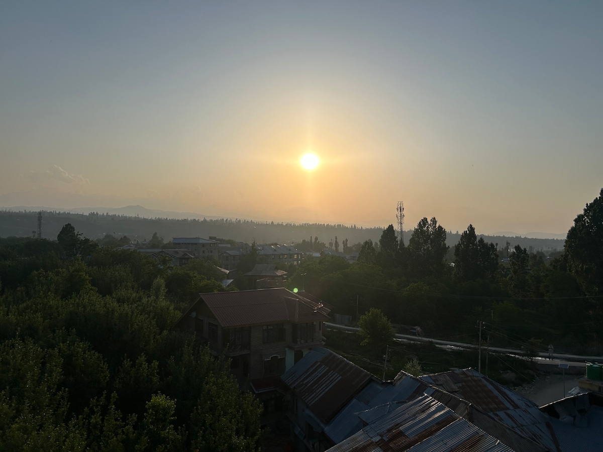 A serene sunset is depicted over a lush green landscape, with the sun slowly descending beyond distant mountains. The scene captures a tranquil atmosphere, with silhouettes of trees and rooftops in the foreground, and the sky displaying warm hues as day transitions to night.