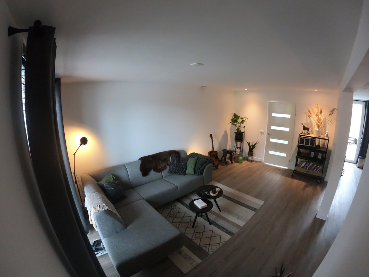 A spacious living room area is showcased, featuring a sectional sofa adorned with a throw blanket. A side table is positioned nearby, accompanied by a floor lamp providing soft lighting. Greenery from potted plants complements the neutral color palette of the walls and flooring.