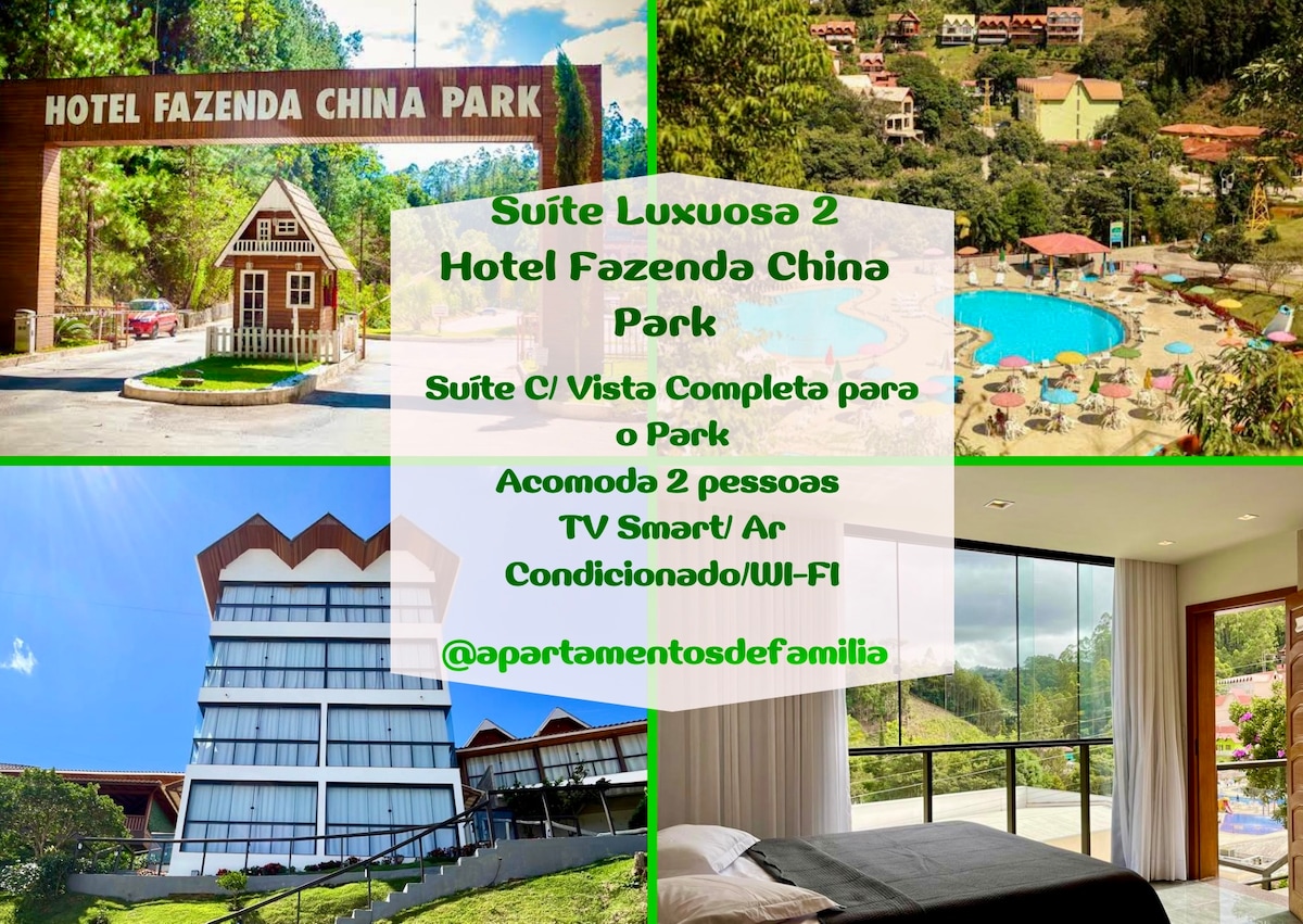An exterior view of Hotel Fazenda China Park highlights the quaint architecture surrounded by lush mountains. The image features a large swimming pool in the foreground, with various attractions visible in the background amid vibrant greenery.