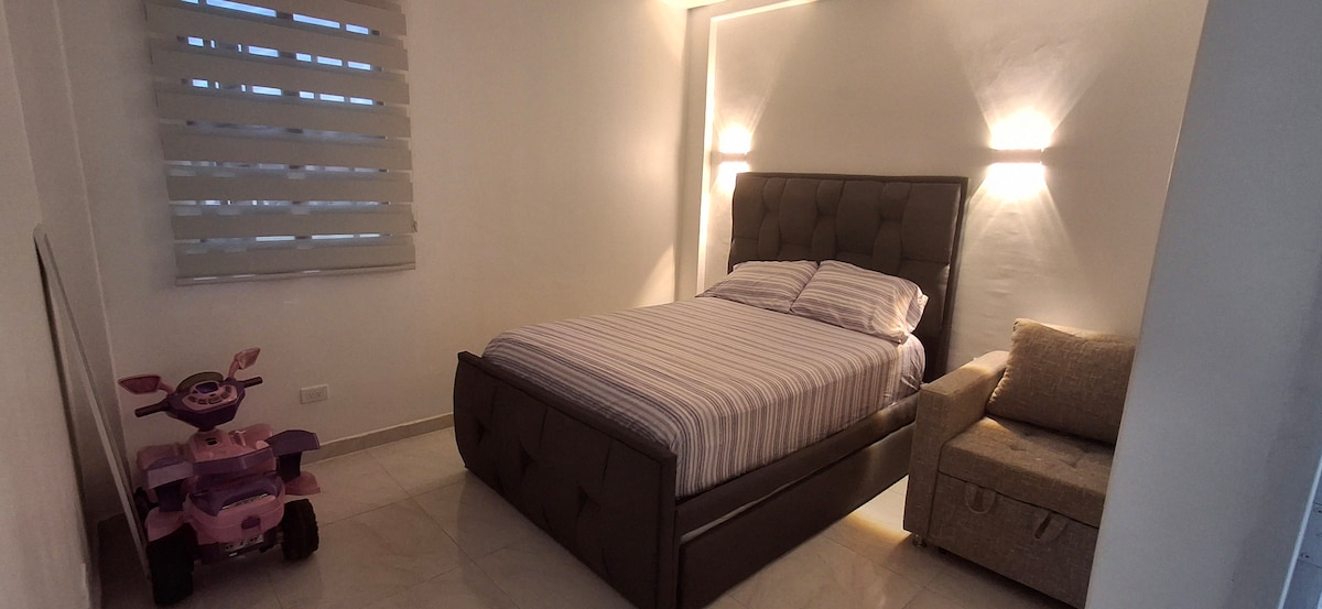 A comfortable sleeping area features a spacious bed adorned with striped bedding. Soft lighting is provided by wall fixtures, creating a serene atmosphere. A cozy armchair is positioned beside the bed, while a children's toy vehicle adds a playful touch to the room.