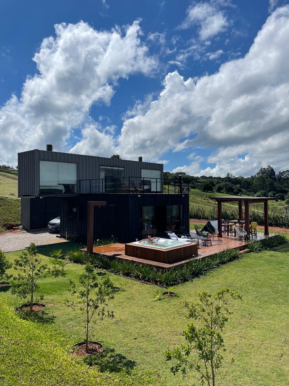 A modern two-story container house is set on a spacious green lawn, surrounded by trees. The exterior features a wooden deck and a jacuzzi, providing a relaxing outdoor space. Bright blue skies and fluffy clouds enhance the tranquil environment.