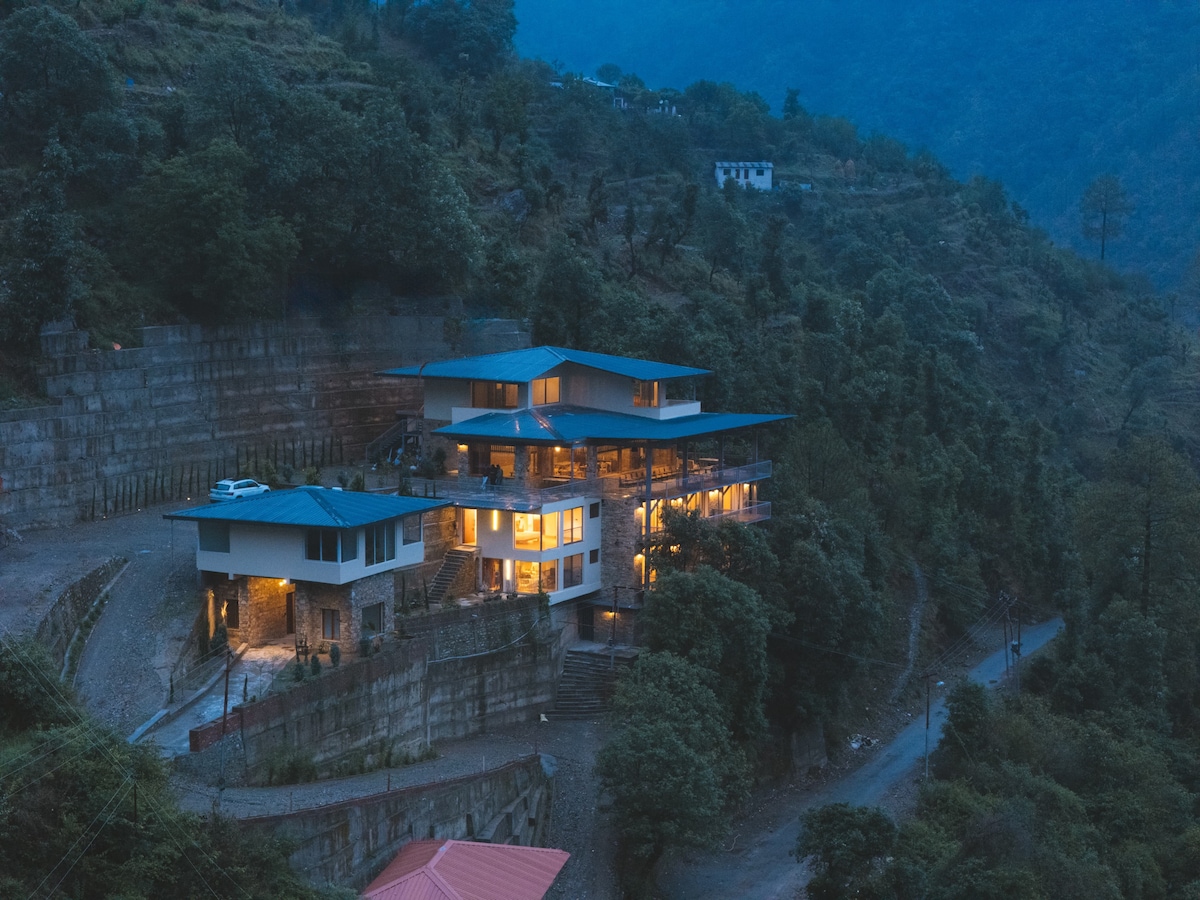 The multi-level structure is nestled in the hillside, presenting a contemporary design with large windows that glow warmly at dusk. Surrounded by lush greenery, the property offers a stunning view of the mountains and includes a winding road leading to its entrance.