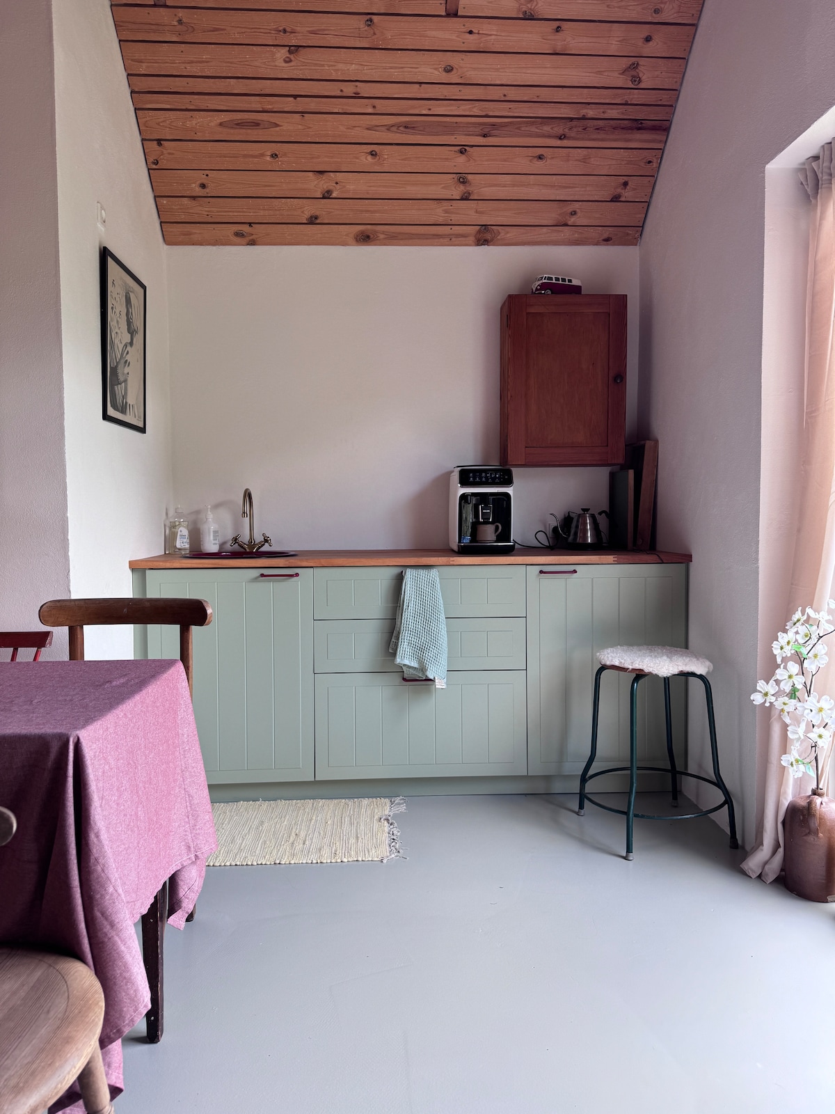 A compact kitchen area is featured, with light green cabinetry and a wooden countertop. Coffee-making appliances are neatly arranged, alongside a sink. A softly colored tablecloth drapes over a dining table, and a small bar stool is positioned nearby, adding to the inviting space.