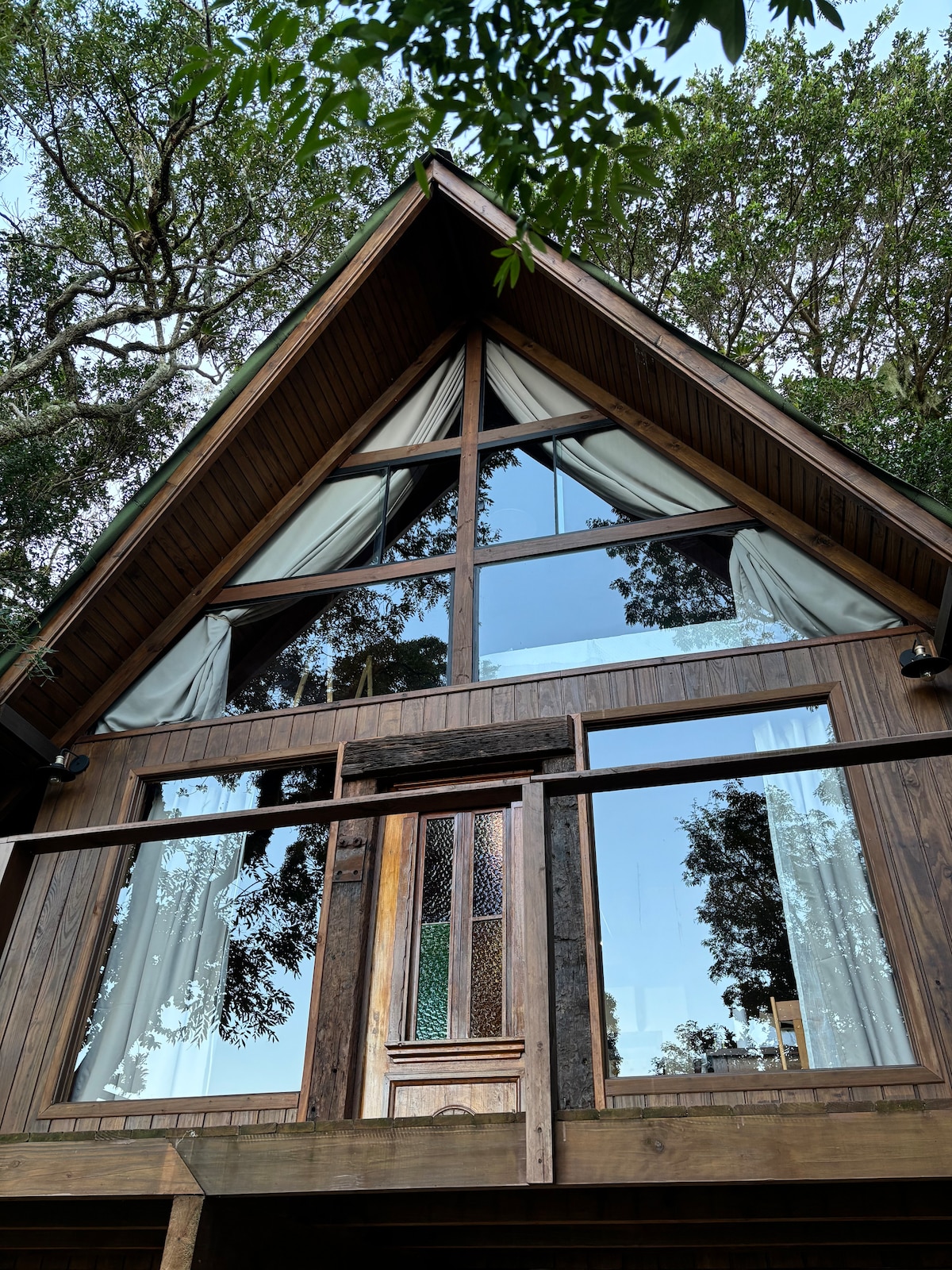The exterior of a stylish A-frame house is showcased, featuring large glass windows that reflect surrounding greenery. Soft curtains are draped at the sides of the windows, and the front entrance is framed with wooden paneling, creating a harmonious blend with the natural environment.