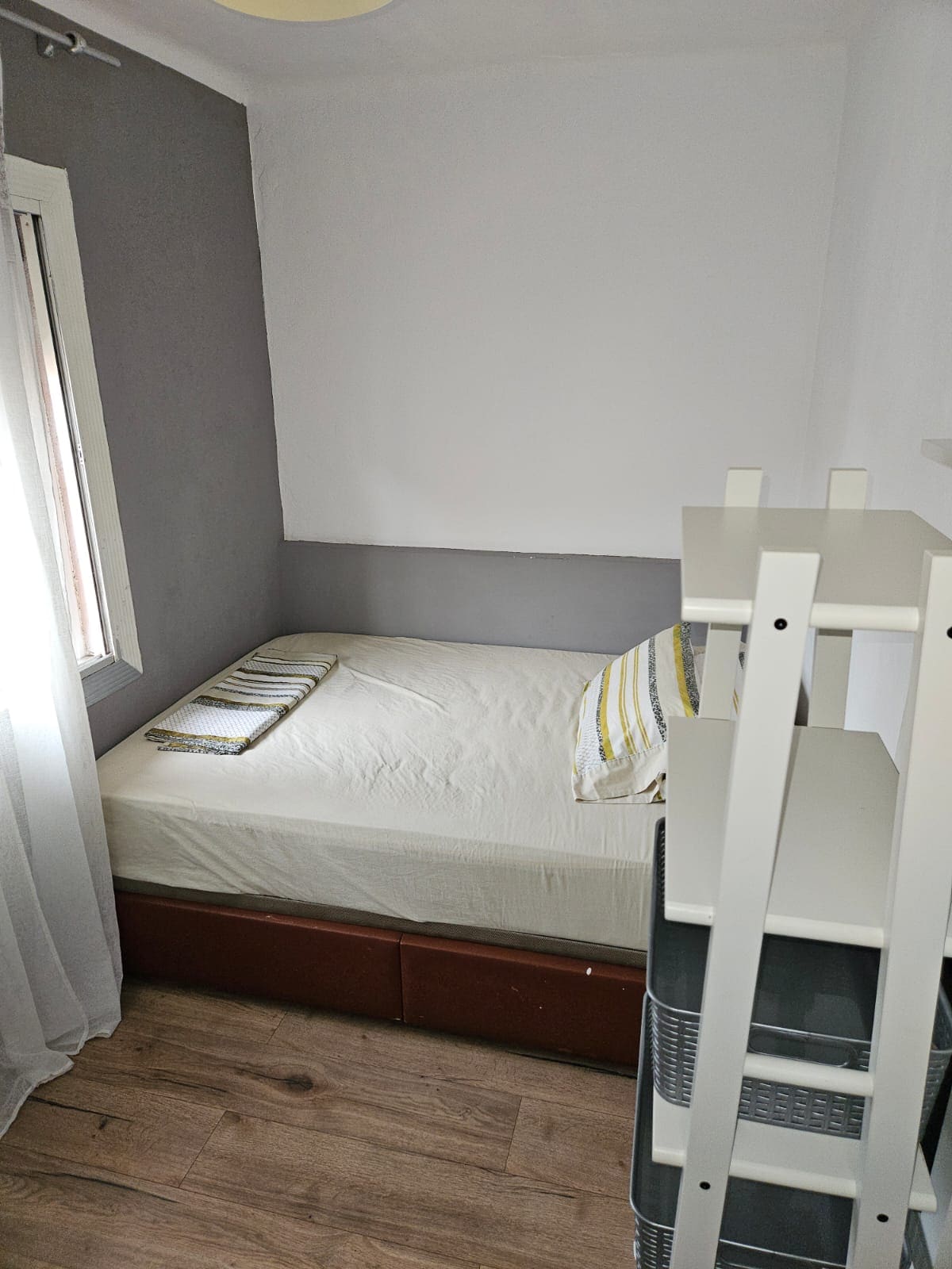 A small bedroom features a single bed with a light-colored duvet and a decorative striped pillow. A window allows natural light to enter, complemented by sheer white curtains. A shelving unit is positioned against the wall, providing storage options.