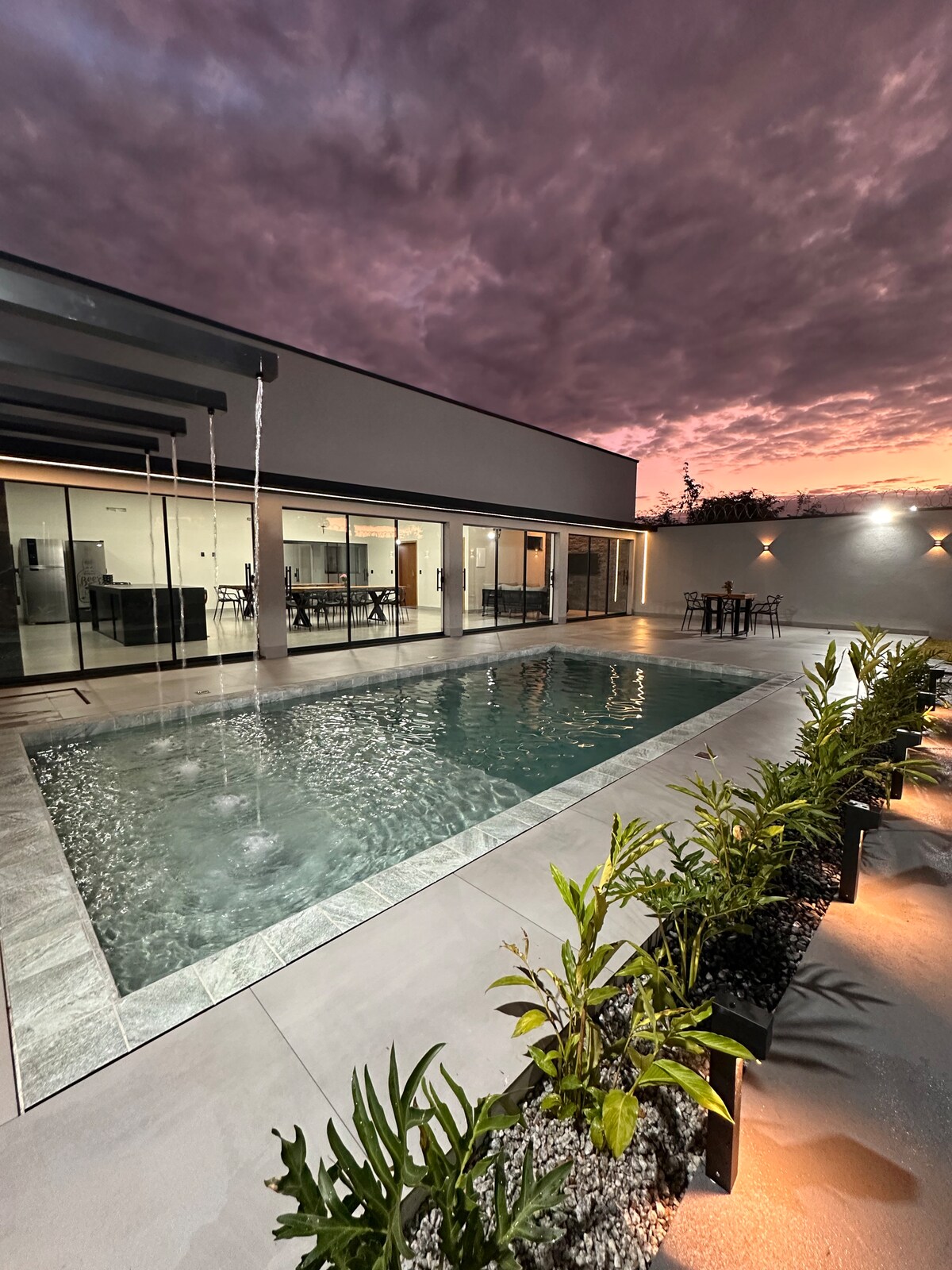 A serene outdoor space features a heated swimming pool, bordered by neatly arranged plants. The evening sky is filled with dramatic clouds, enhancing the tranquil setting. Glass doors connect the pool area to an indoor space, creating a seamless transition between outdoor and indoor living.