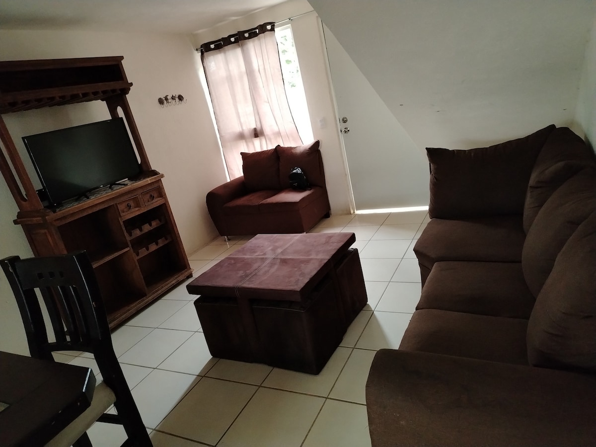 A cozy living area is furnished with a large brown sofa and a smaller armchair. A wooden TV stand displays a television and provides shelving. A square coffee table is positioned centrally, with tile flooring and natural light filtering through the window covered by sheer curtains.