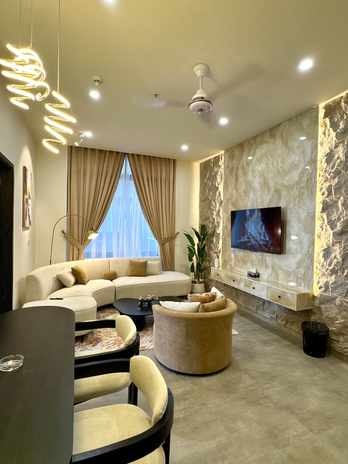 The living room features a curved sectional sofa accompanied by a round chair, complemented by a glass coffee table. A media unit with a wall-mounted television is adorned by a striking stone wall. Natural light filters through large windows, enhanced by soft curtains.