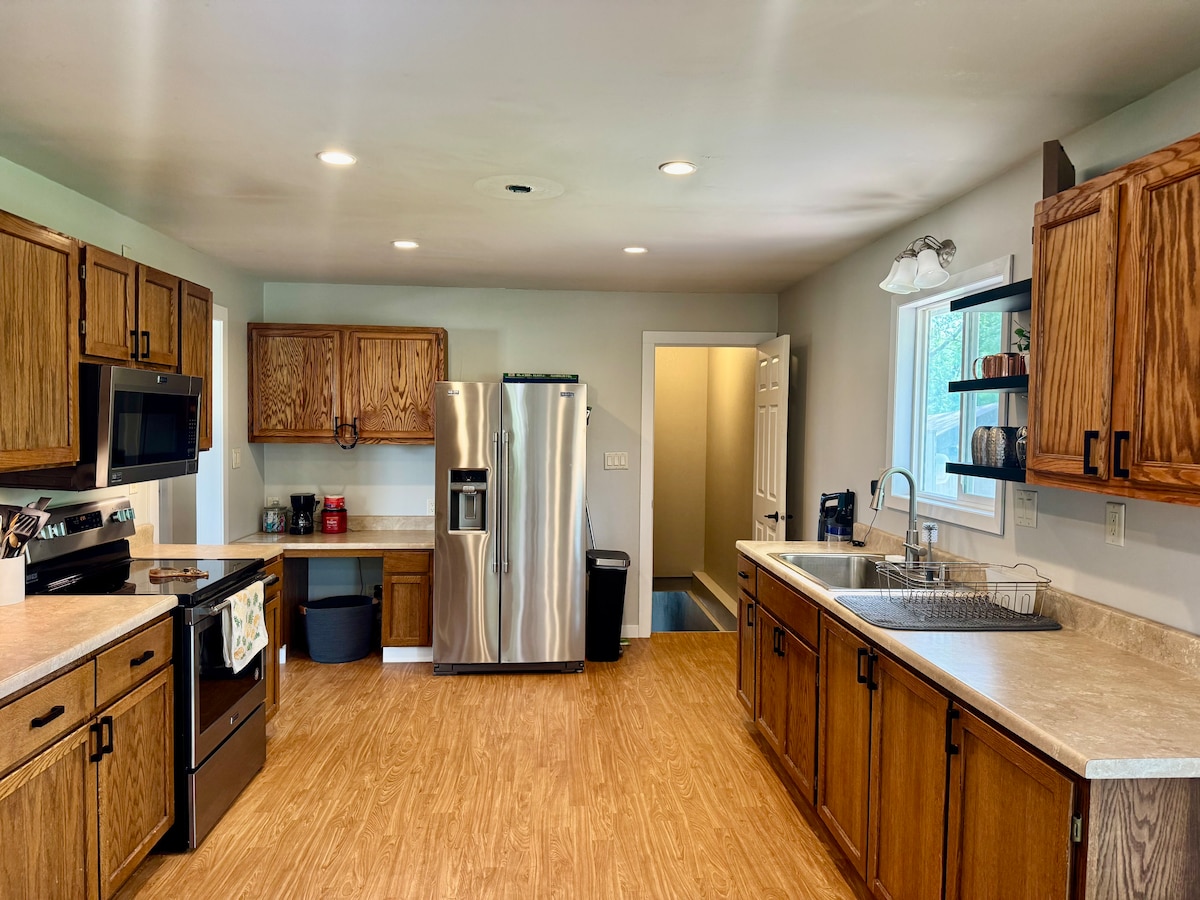 A spacious kitchen features wooden cabinetry and modern appliances. A large stainless steel refrigerator stands beside an oven and a microwave. A double sink is located beside a window, while ample counter space is available for preparation. Access to a nearby pantry is provided.
