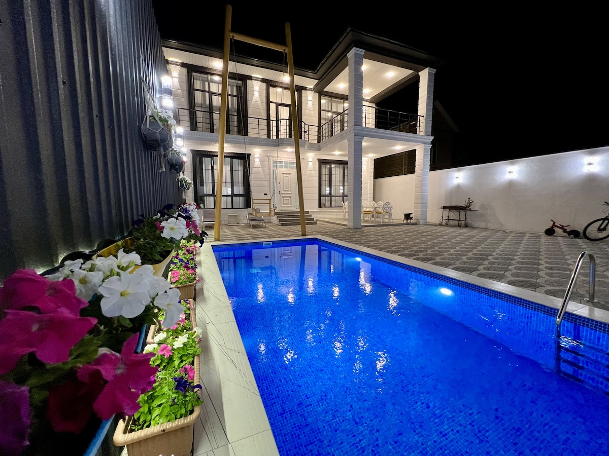 A well-lit outdoor area features a sparkling blue pool, surrounded by colorful flower arrangements in planters. The two-story villa is visible in the background, with spacious terraces and elegant lighting illuminating the entrance.