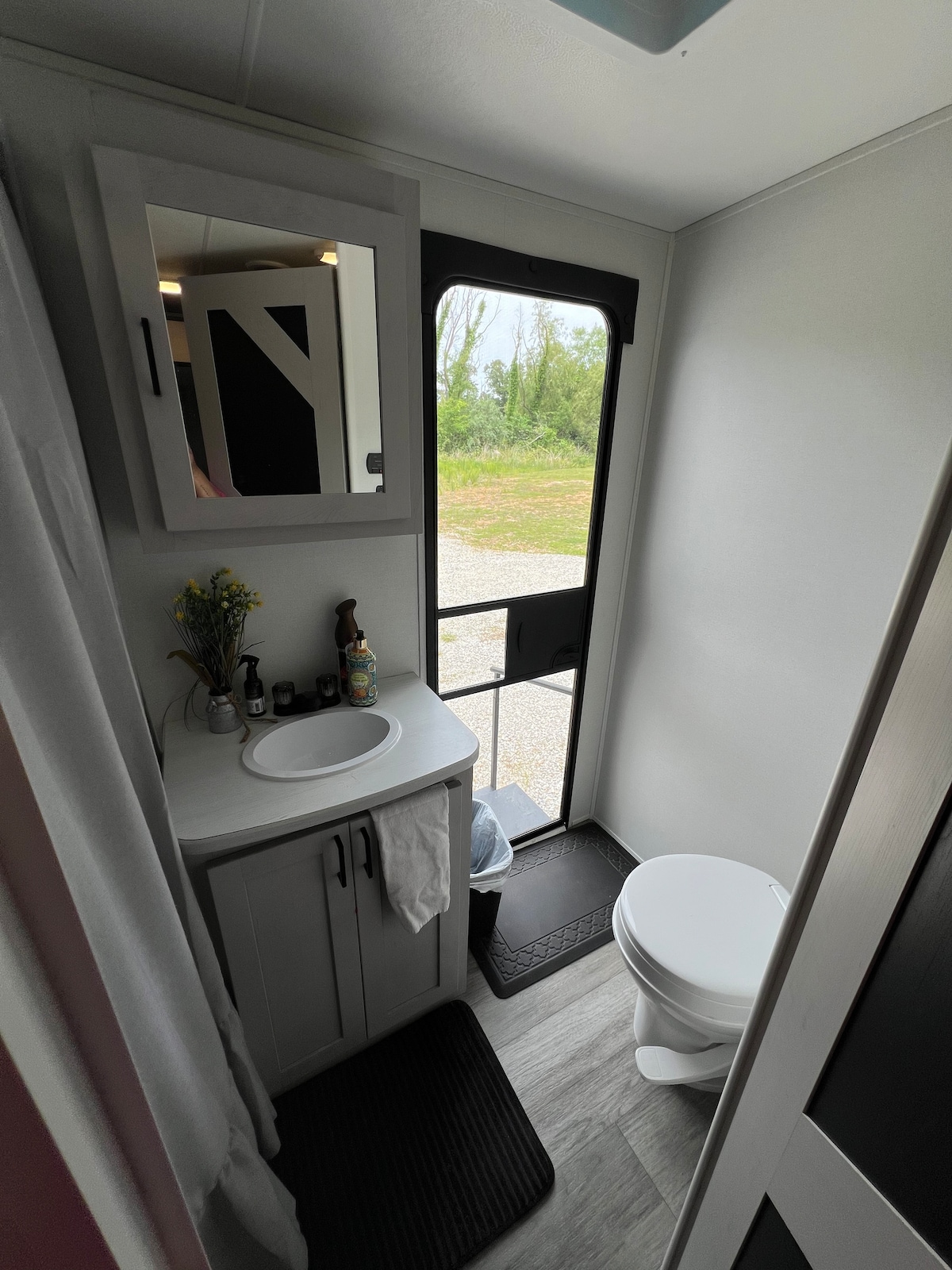 A compact bathroom space features a white toilet and a small sink with a mirror above it. Natural light enters through a large window, and a mat is placed on the floor. A small shelf holds toiletries, contributing to a clean and functional atmosphere.