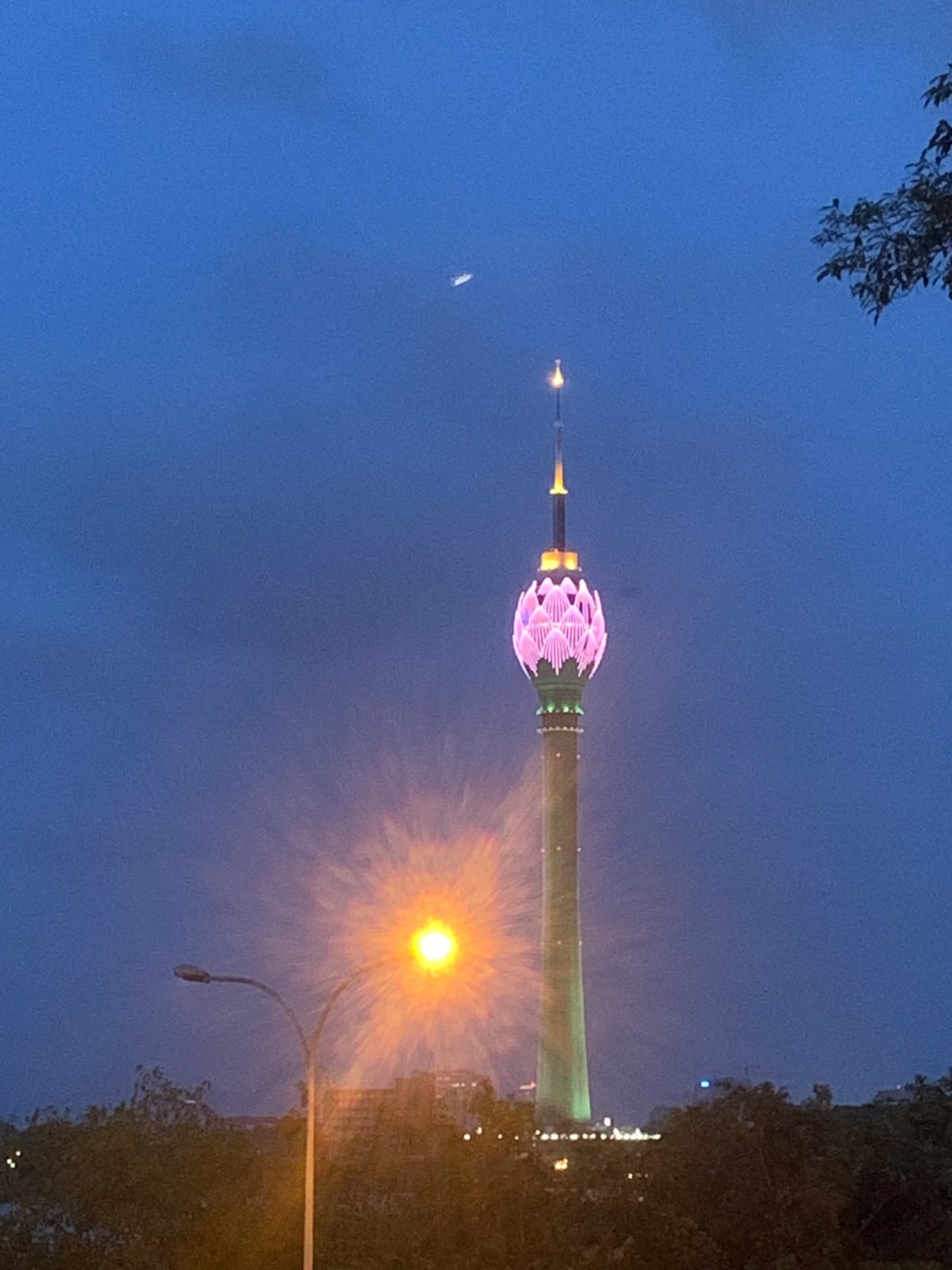 A tall tower is illuminated in vibrant purple and green lights against a dusky blue sky. A burst of light emanates from a nearby street lamp, adding to the evening ambiance. The silhouette of the cityscape is visible at the base of the tower.