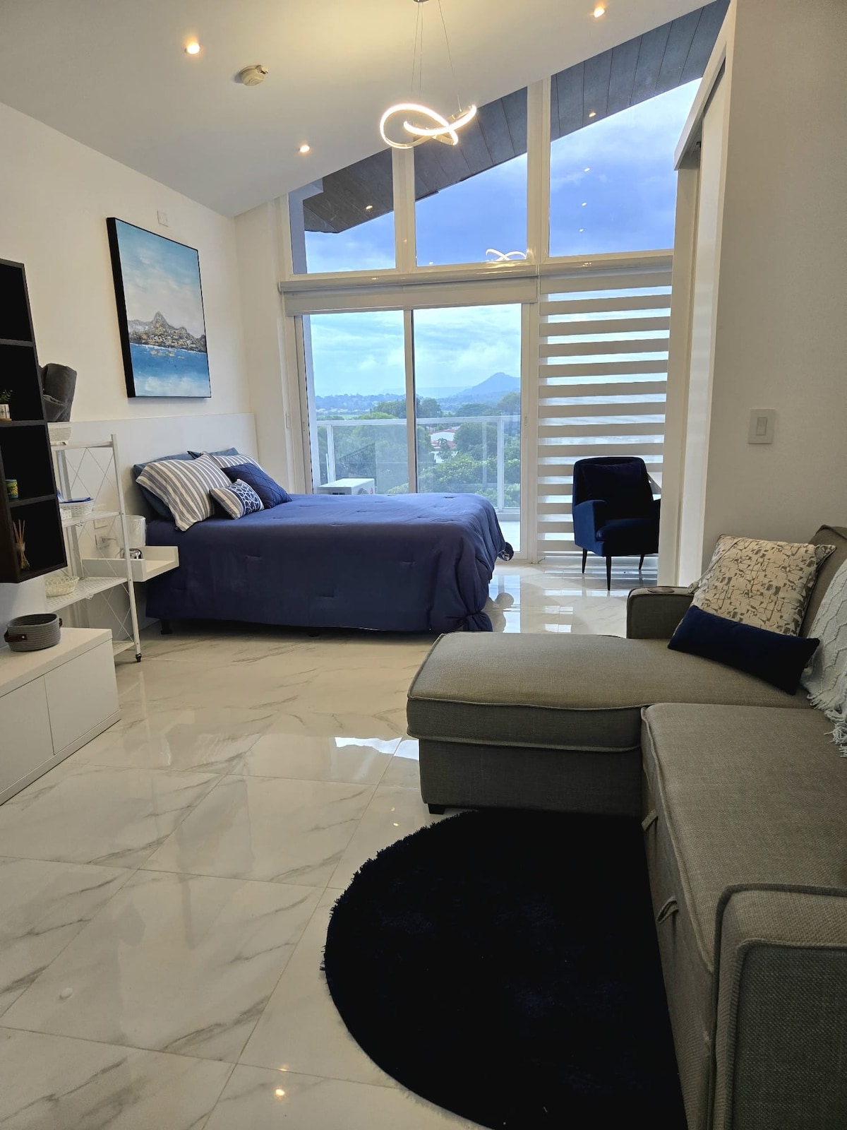 A modern bedroom is showcased with a spacious bed featuring a blue blanket. Floor-to-ceiling windows allow natural light to flood the room, revealing a view of the mountains. A cozy seating area includes a gray couch and a dark rug, creating a comfortable environment.