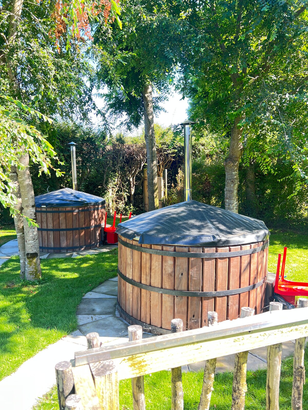 Two wooden hot tubs are nestled among lush greenery, surrounded by tall trees. A stone pathway leads to the tubs, which are positioned on a manicured lawn. Red chairs are seen nearby, enhancing the natural setting.